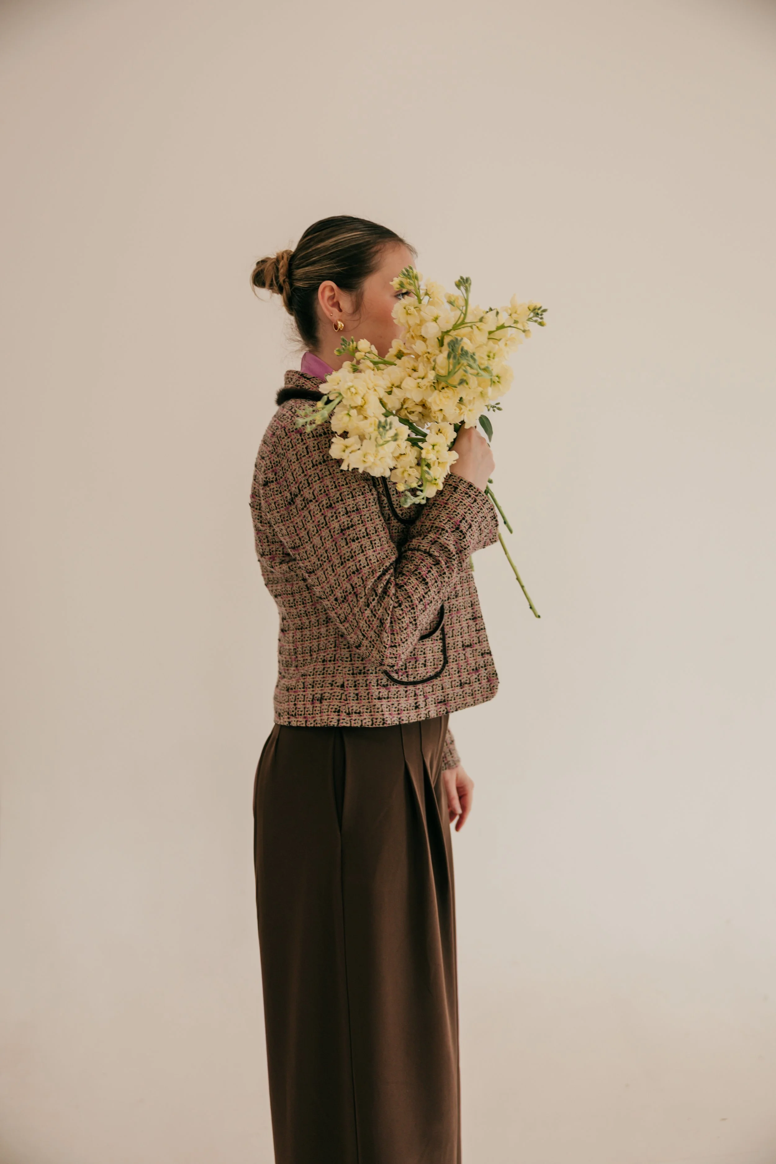A woman holding a large bouquet of yellow flowers, standing sideways against a plain white background, wearing a patterned jacket and brown pants.