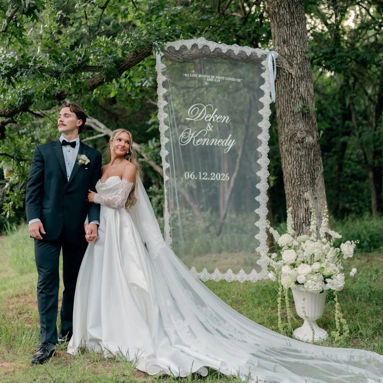 A bride and groom holding hands outdoors next to a large transparent wedding sign with floral decorations and the wedding date. Beautiful white floral arrangement in an urn next to a couple.