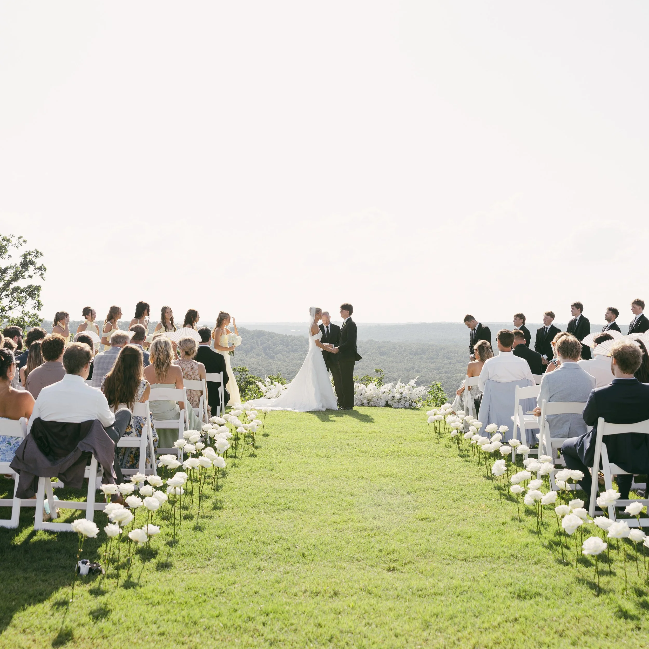 Outdoor wedding ceremony with a bride and groom exchanging vows under a bright sky, surrounded by seated guests on a grassy lawn decorated with white flowers, with trees in the background. All rose aisle with white roses sticking up from the ground