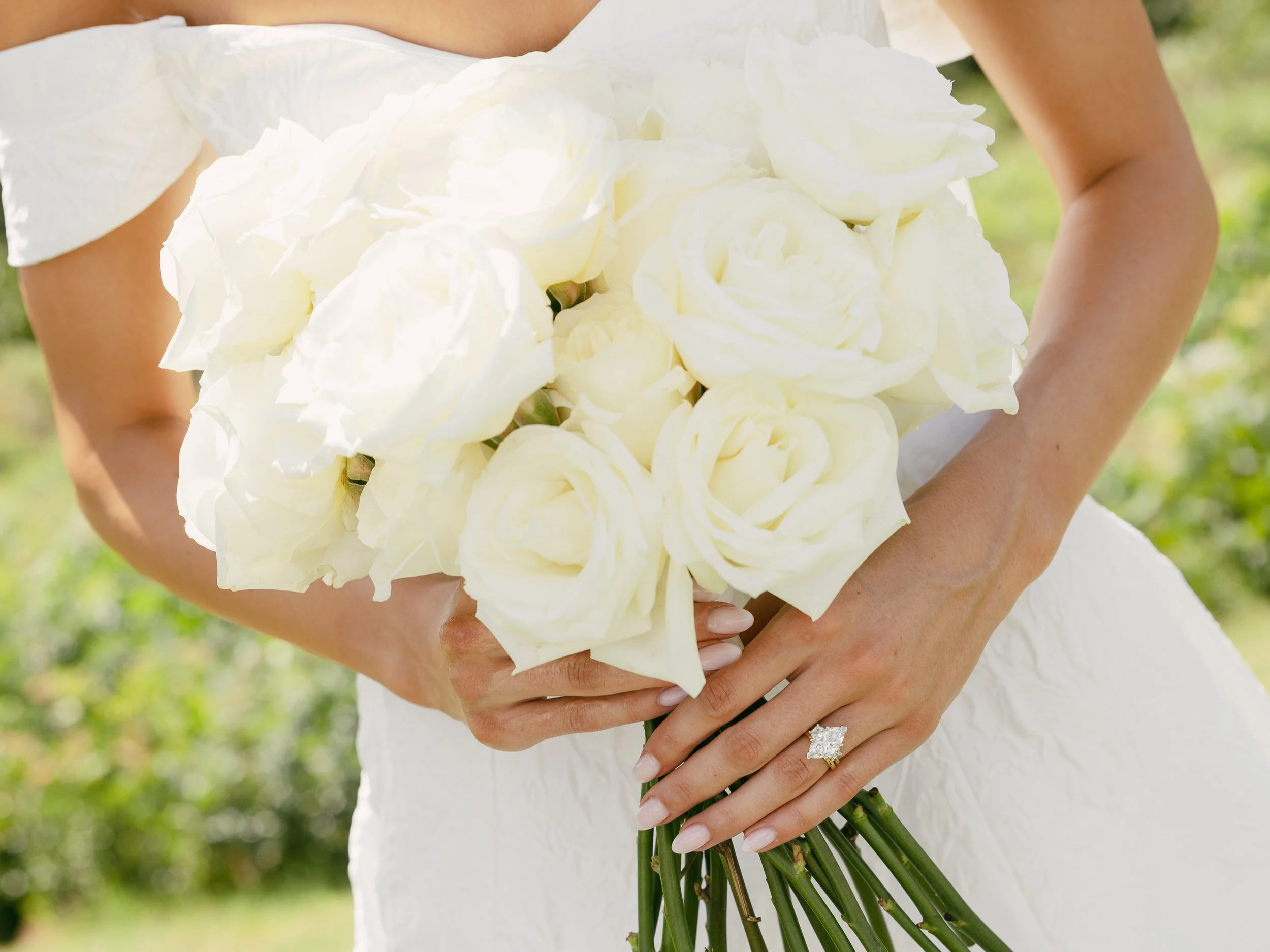 A woman wearing a white dress holds a large bouquet of white roses outdoors.
