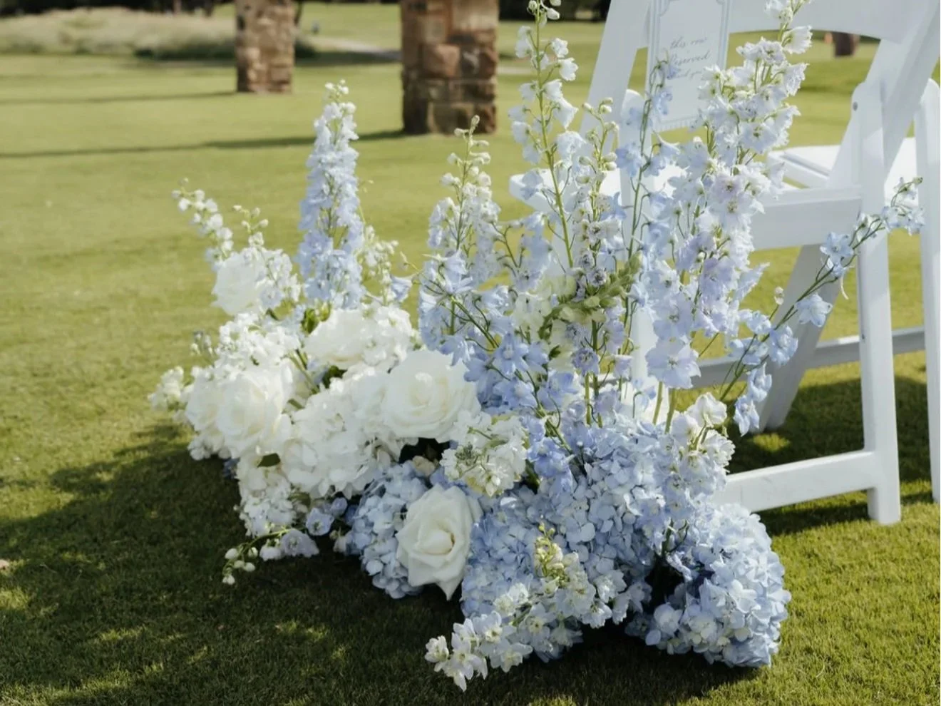 Arrangement of white roses and blue delphiniums on grass, with white chairs in the background.