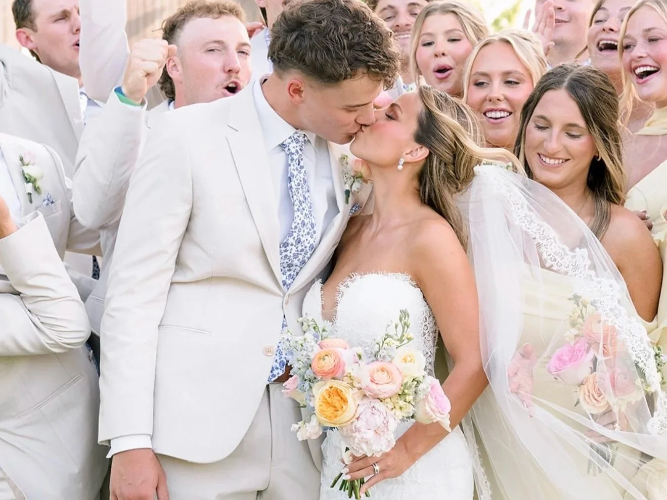 A newlywed couple shares a kiss at their wedding, surrounded by smiling guests, with the bride holding a bouquet of pink and yellow roses.