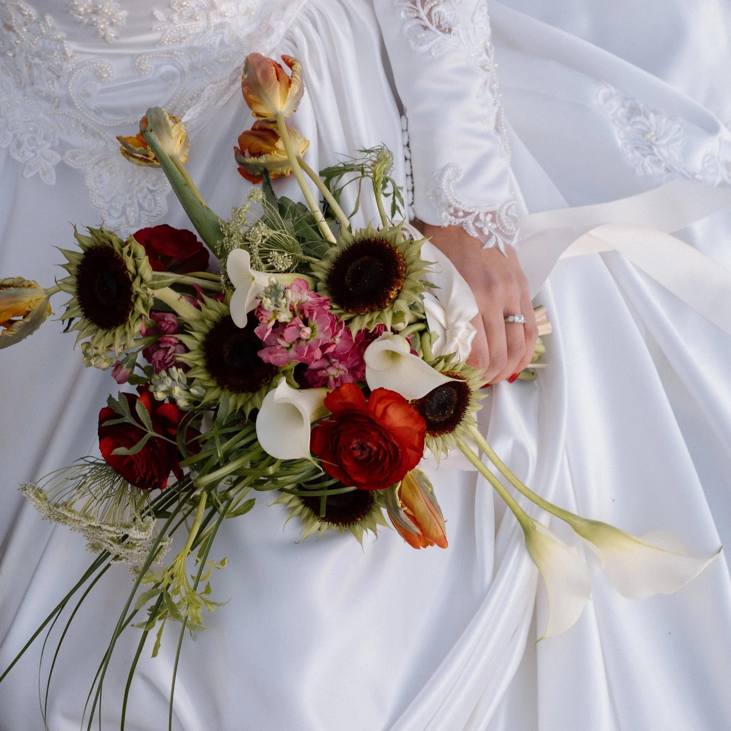 A bridal bouquet with sunflowers, calla lilies, roses, and other flowers resting on a white wedding dress with lace details, held by a bride with a ring on her finger.