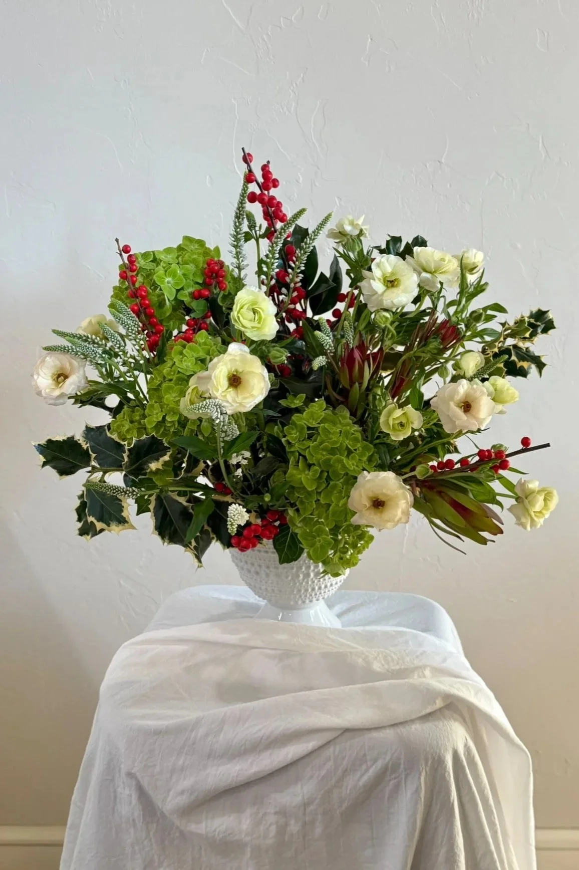 A festive holiday flower arrangement in a white textured vase on a draped white cloth against a plain wall. The arrangement includes white flowers, green foliage, red berries, and sprigs of holly.