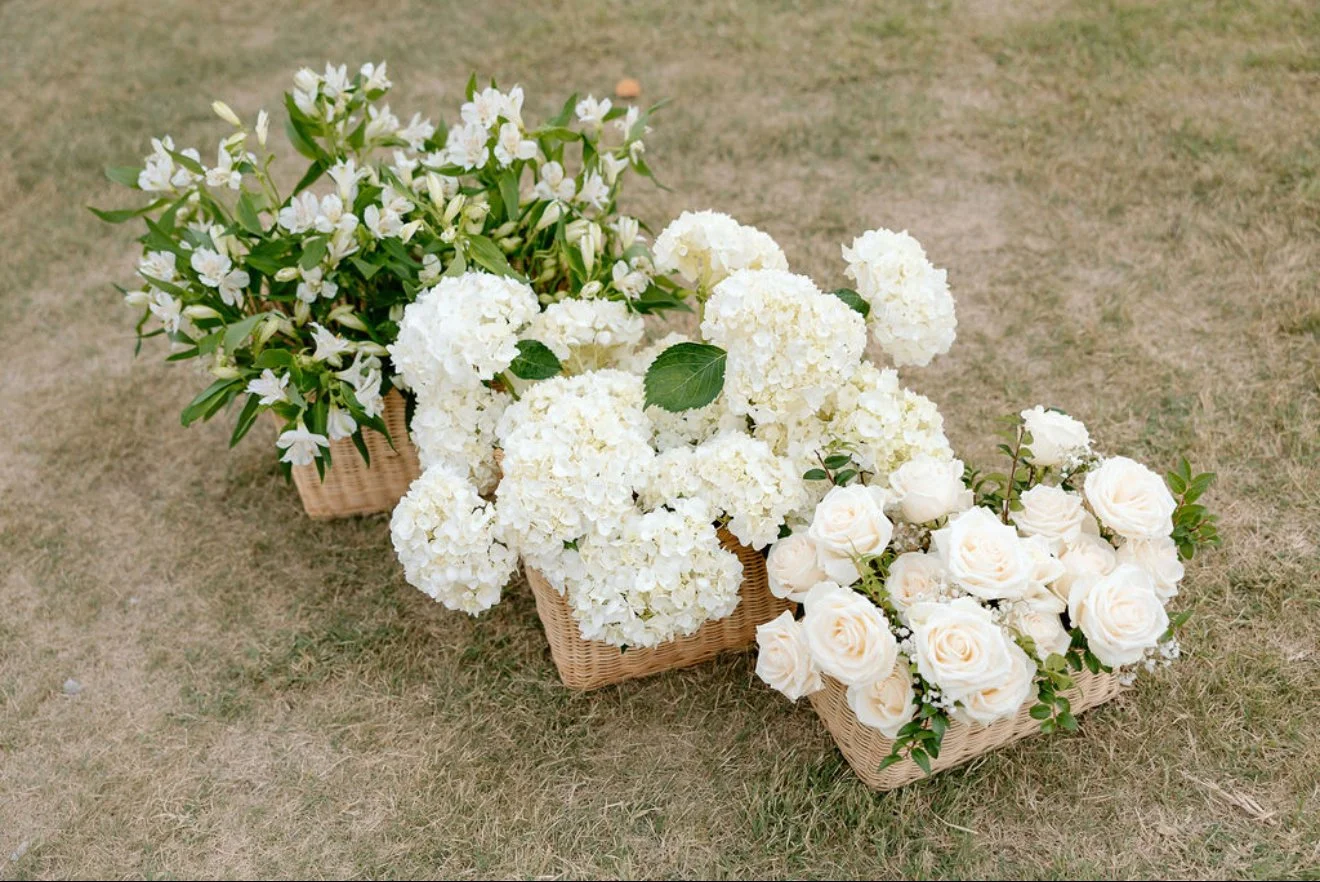 Three wicker baskets filled with white floral arrangements, including hydrangeas, roses, and other white flowers, placed on a grassy outdoor area.