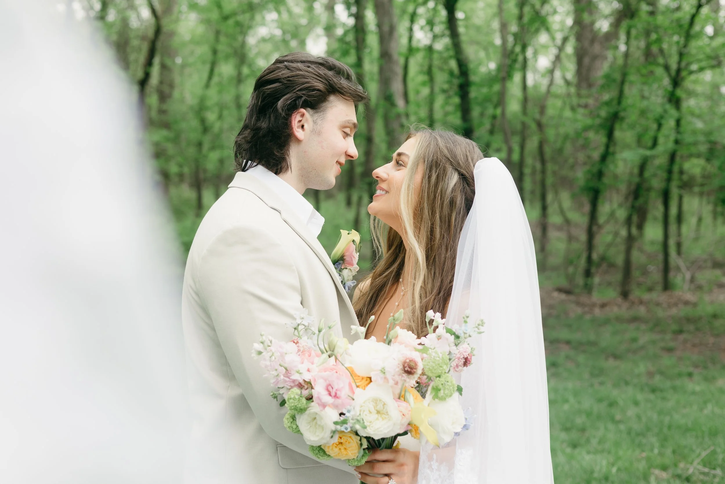 Bride and groom standing close together outdoors, holding a bouquet of flowers, gazing at each other, with a background of green trees.