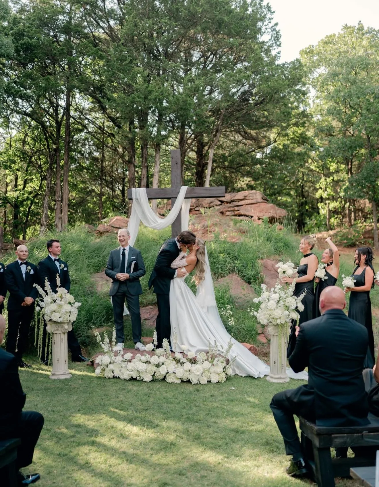 A wedding ceremony outdoors with a couple kissing in front of a cross, surrounded by friends and family dressed in formal attire, with greenery and trees in the background. A lush and fluffy white floral meadow and pillar arrangements at the alter.
