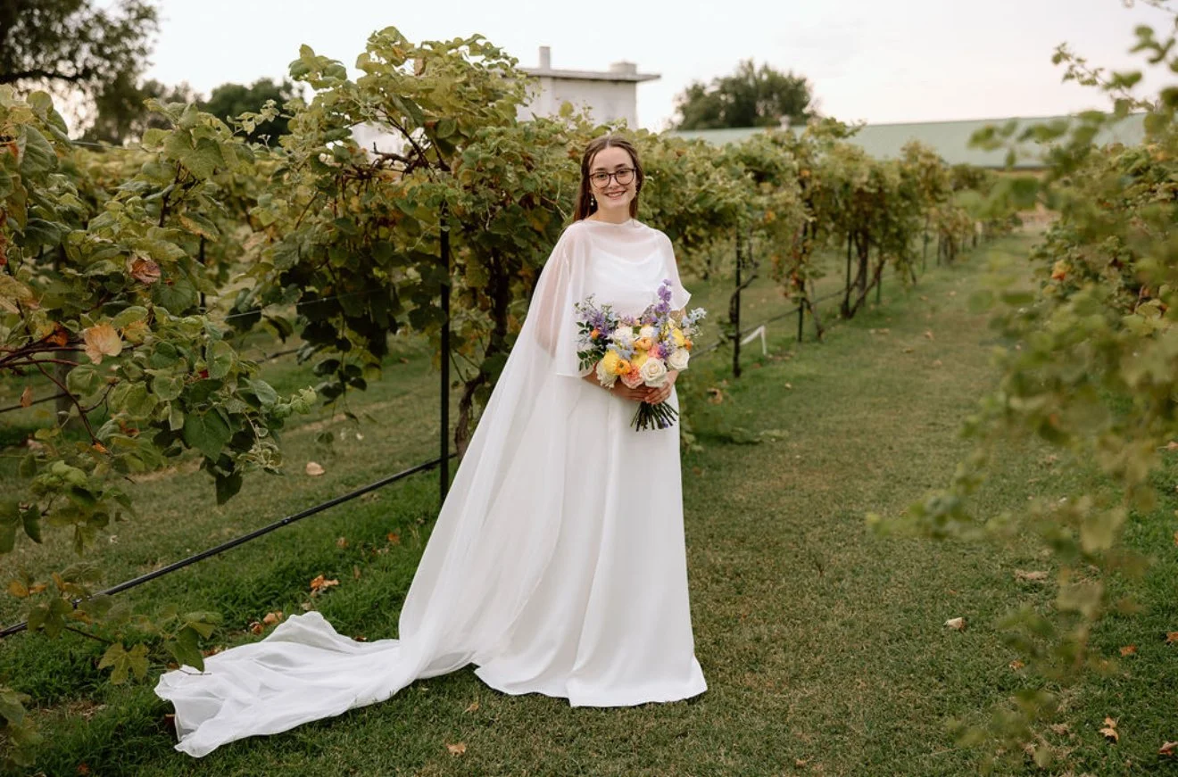 A young woman in a white wedding dress holding a colorful bouquet standing in a vineyard.