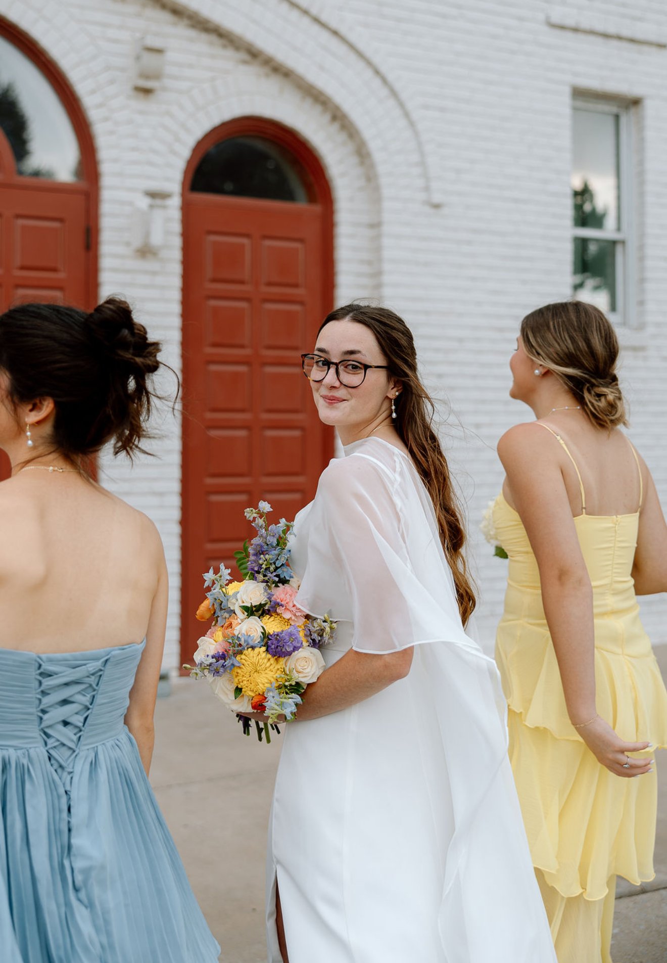 A bride holding a bouquet of colorful flowers smiles and turns her head back as she stands with bridesmaids outside a white brick building with red doors.