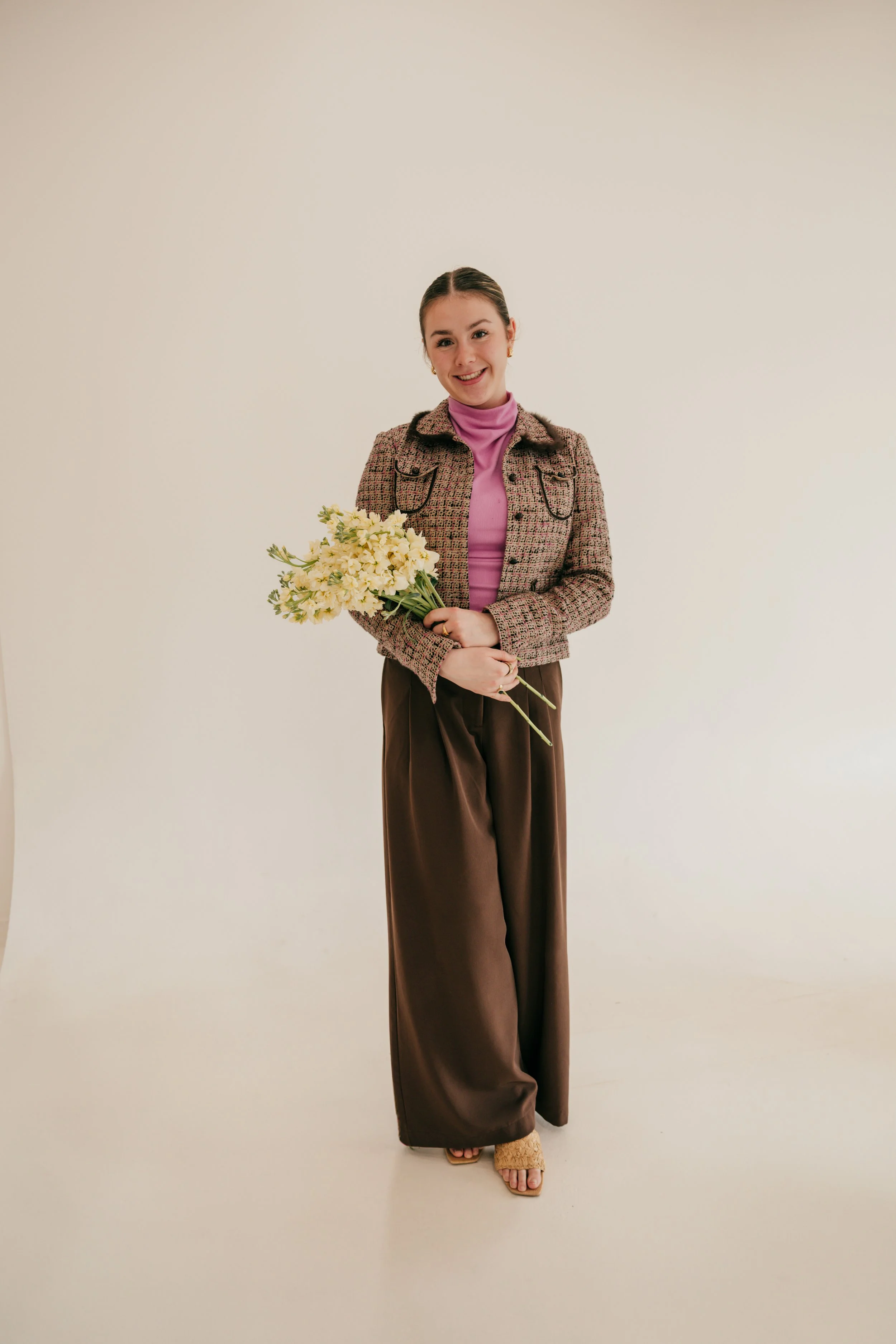 A woman standing against a plain light-colored background, smiling, holding a bouquet of yellow and white flowers, wearing a pink turtleneck, a checkered blazer, wide brown pants, and sandals.