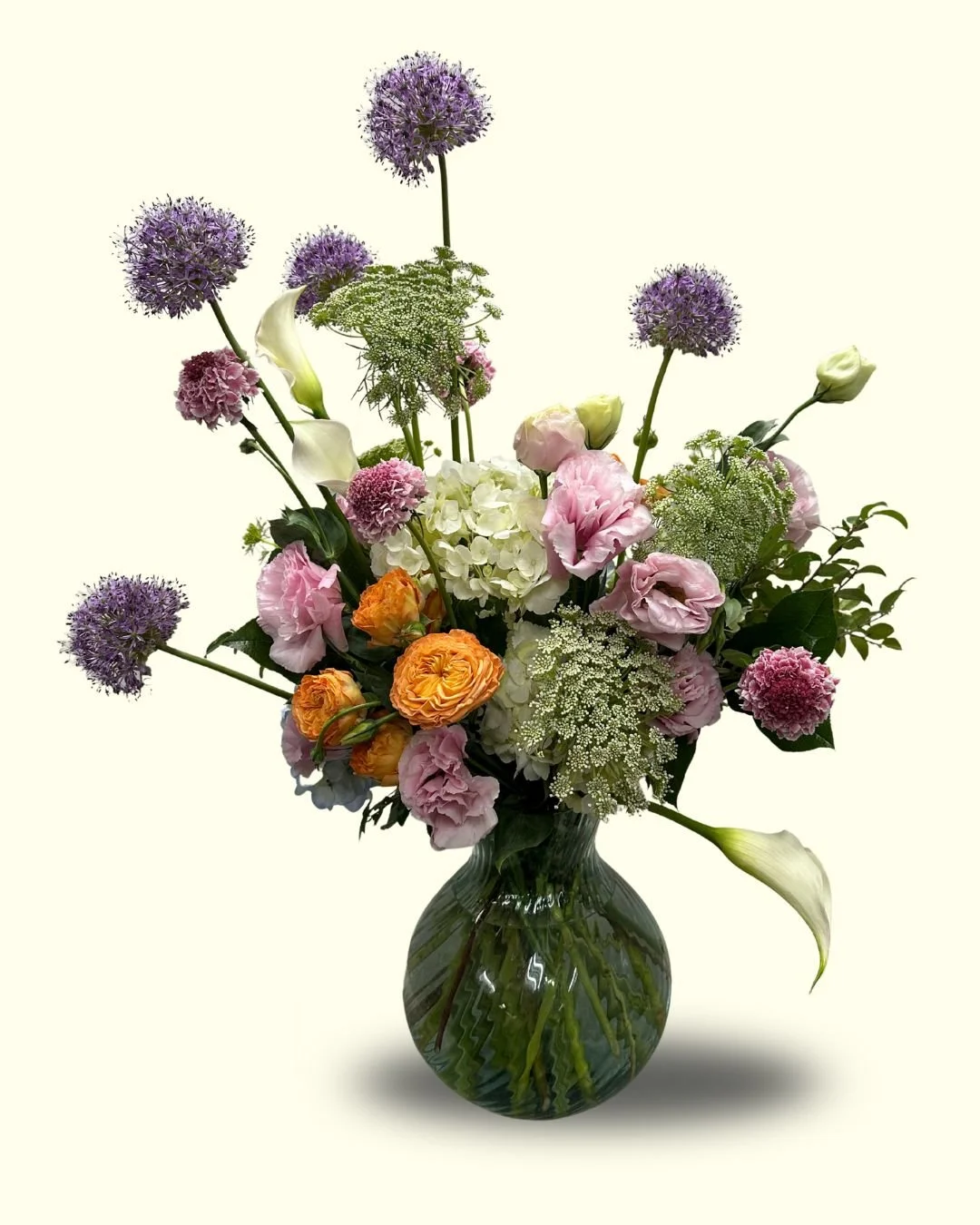 A colorful bouquet of flowers in a green glass vase, including purple alliums, pink carnations, white hydrangeas, orange ranunculus, calla lilies, and other greenery, set against a plain light background.