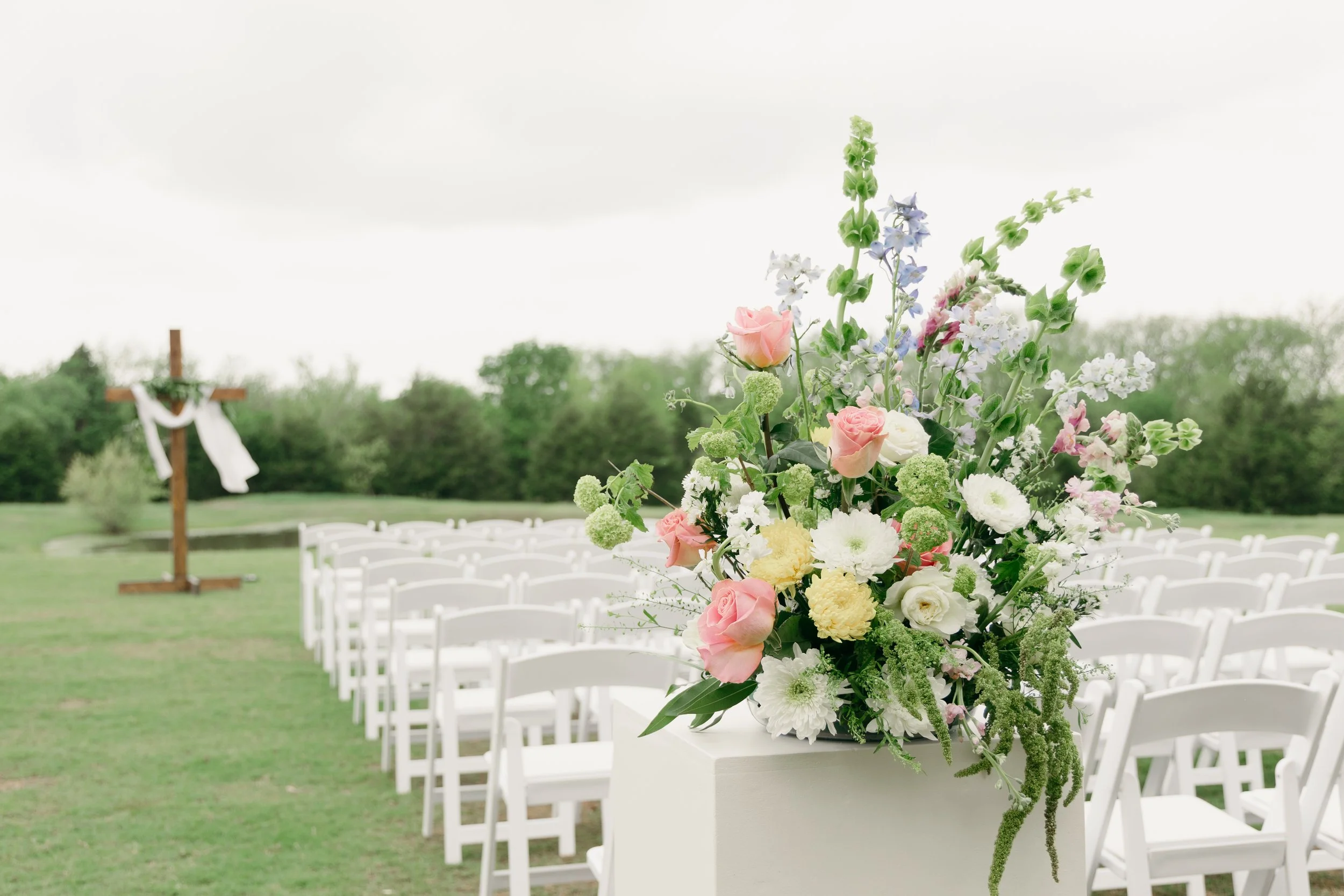 Flower arrangement with pink roses, white and yellow flowers, and greenery on a pedestal at an outdoor wedding ceremony with white chairs and a cross in the background
