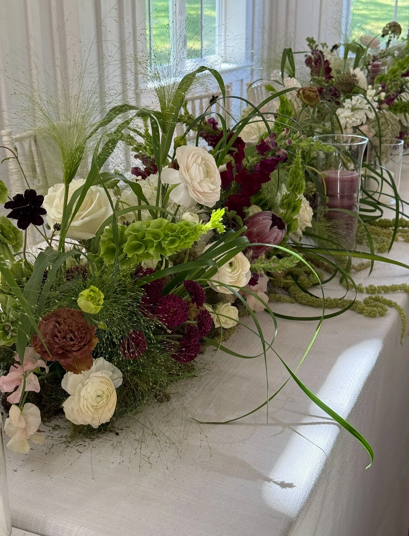 A floral arrangement with white, purple, and brown flowers and green foliage on a white table near a window. Textural and elevated greens on a lush floral head table.