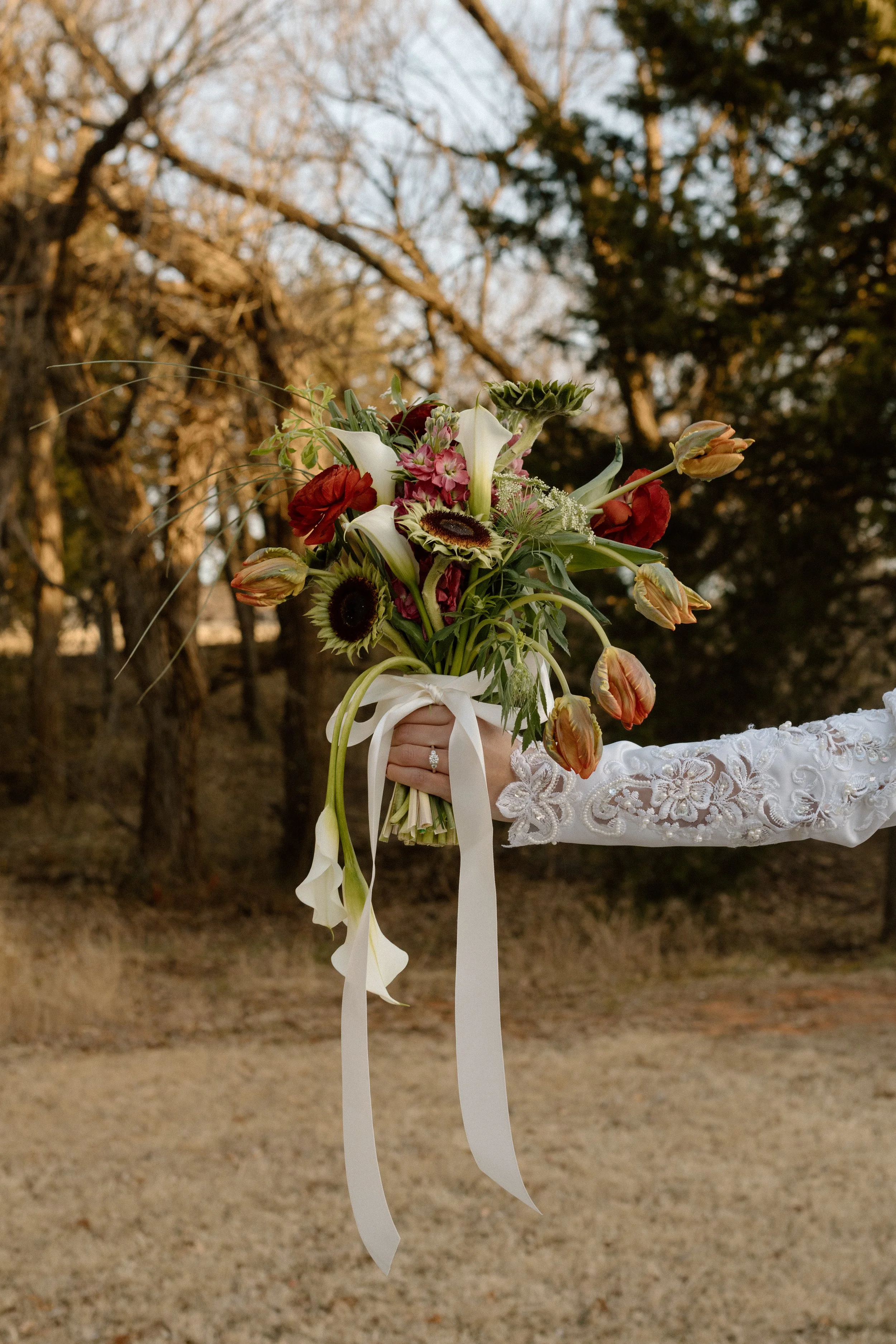 A person in a lace sleeve holding a bouquet of various flowers outdoors with trees in the background.