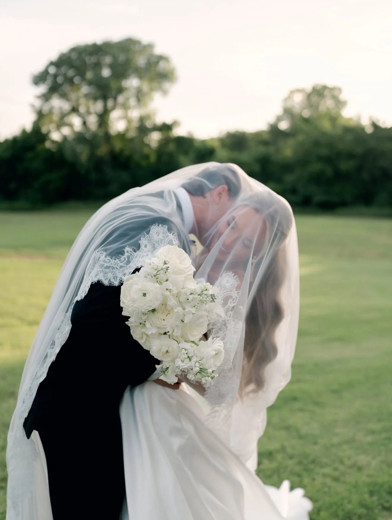 A bride and groom embrace outdoors during a wedding, with the bride wearing a lace wedding dress and veil, holding a bouquet of white flowers, and the groom in a dark suit, with trees in the background.