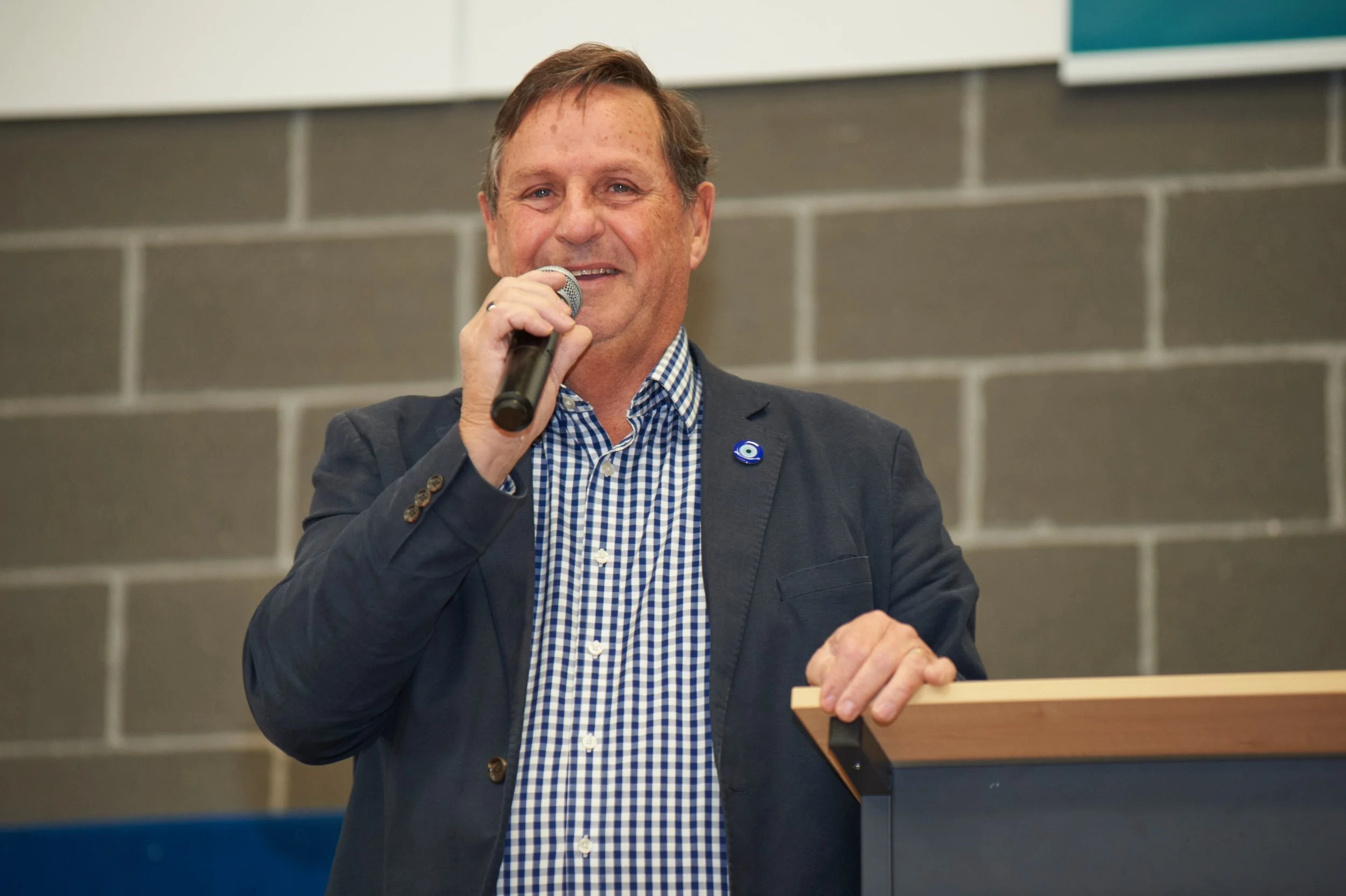 A middle-aged man is speaking into a microphone at a podium in an indoor setting with a gray brick wall in the background. He is wearing a checkered shirt with a dark blazer and has a small round blue pin on his blazer lapel.