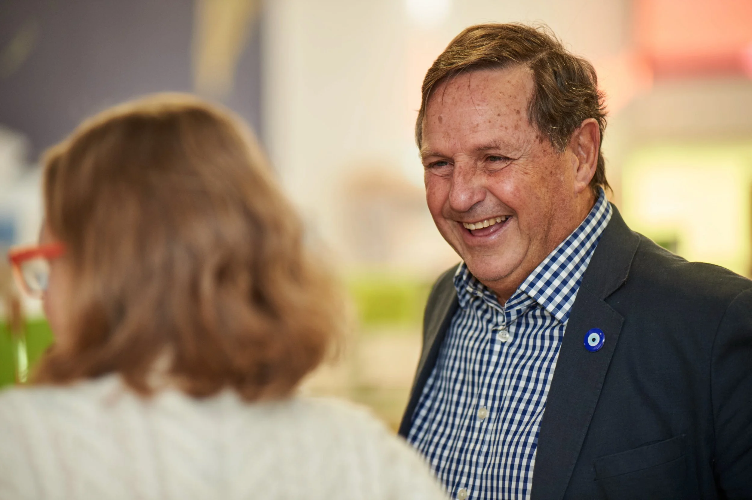 A man with a checkered shirt and a dark blazer smiling and talking to a woman with glasses in an indoor setting.