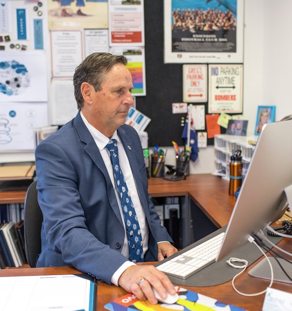 A man in a blue suit and tie working at a desk with a computer in an office decorated with posters, signs, and office supplies.