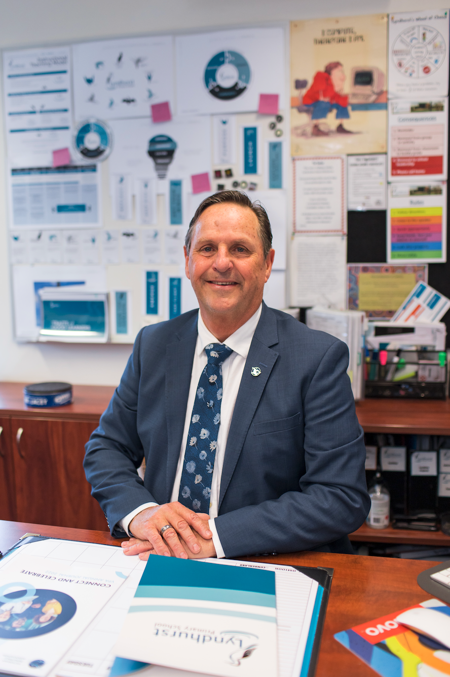 A professional man in a suit sitting at a desk with a smile, in an office with various charts and posters on the whiteboard behind him.