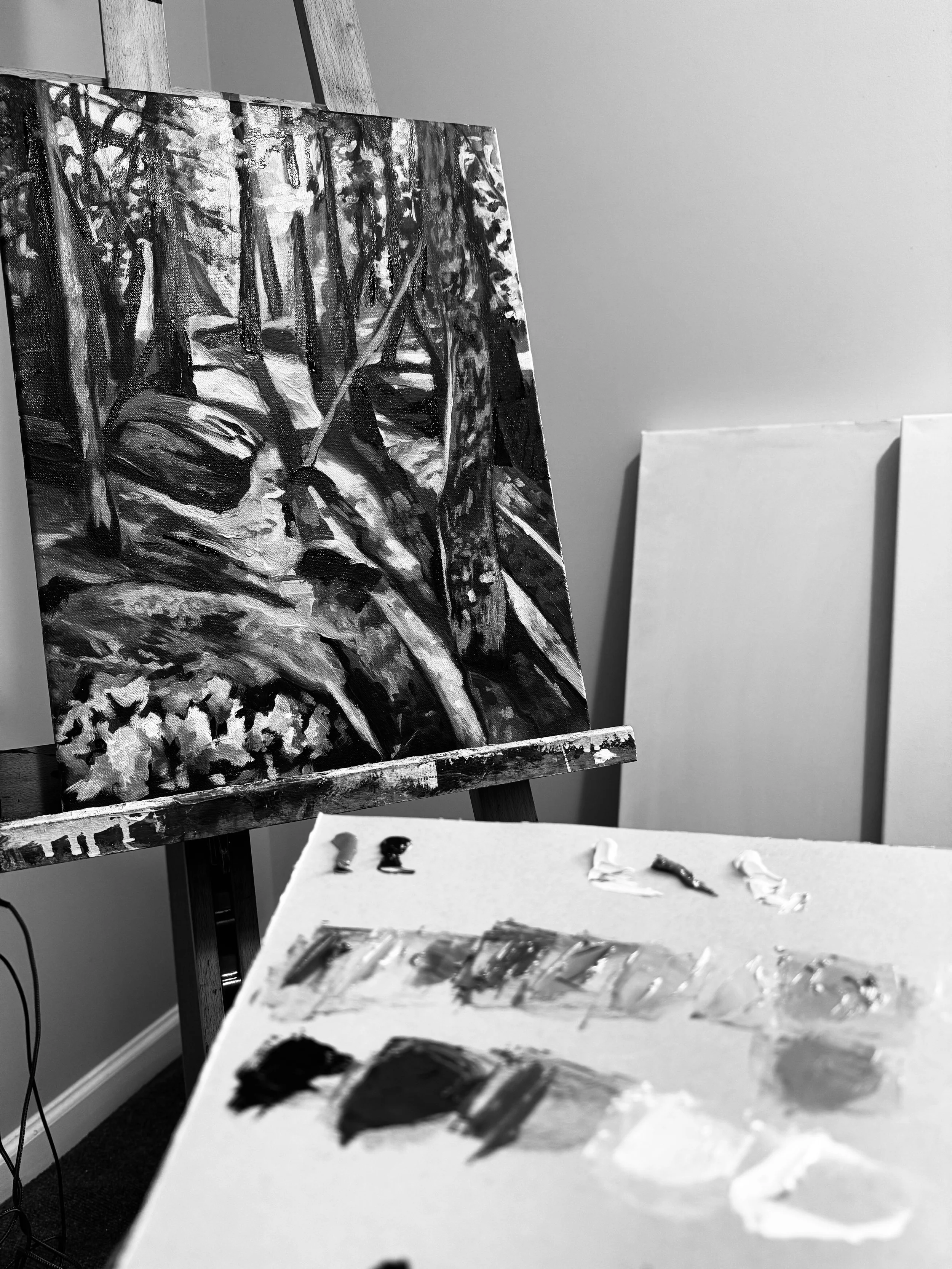 Black and white photograph of a painting in progress depicting a forest scene with trees and rocks, with a palette and brushes in the foreground.