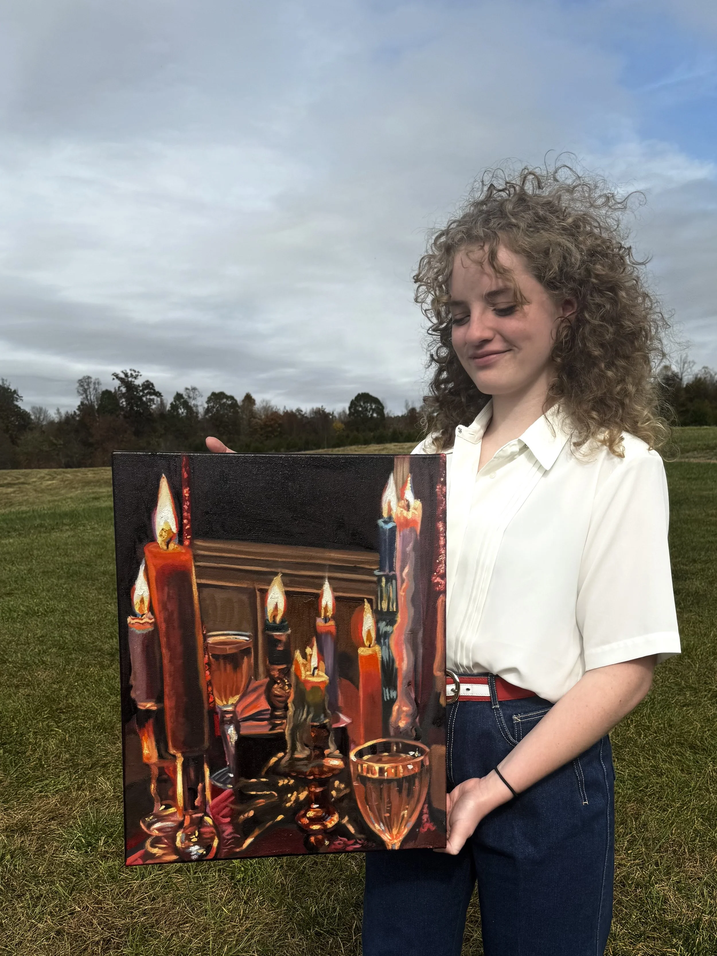 Young woman standing outdoors on a grassy field, holding a colorful painting of burning candles and glasses, with a cloudy sky and trees in the background.