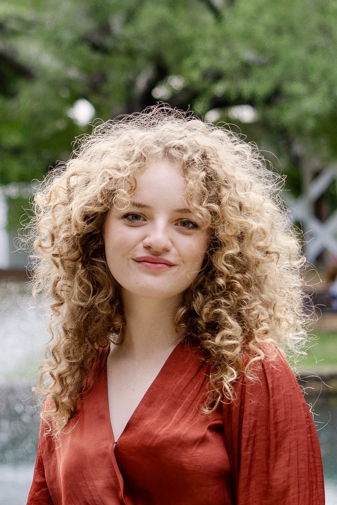 A woman with curly blonde hair wearing a red top, standing outdoors with trees and a water feature in the background.