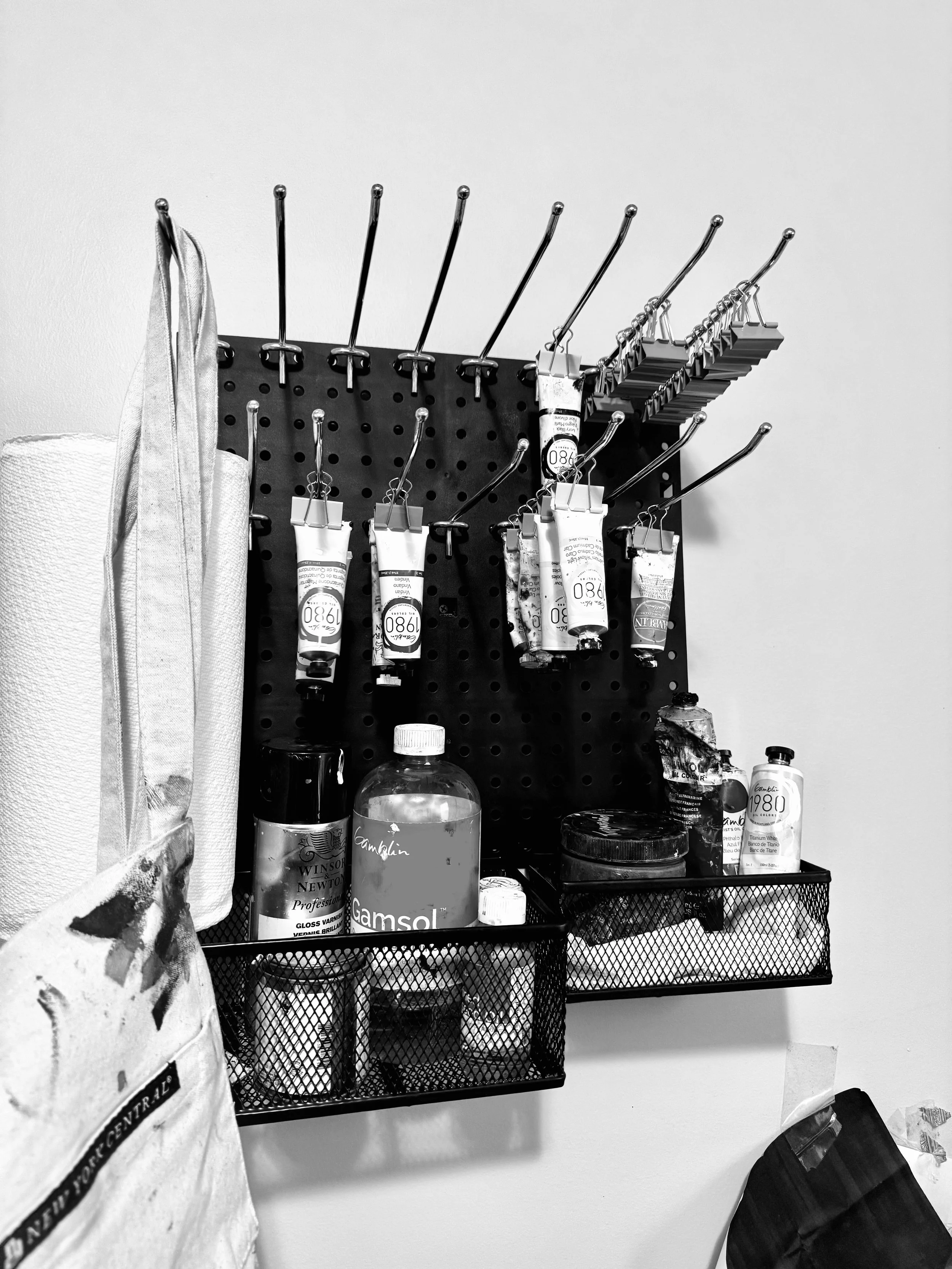 A black pegboard with various art supplies including paint tubes, brushes, and containers, and a mesh tray holding additional supplies.