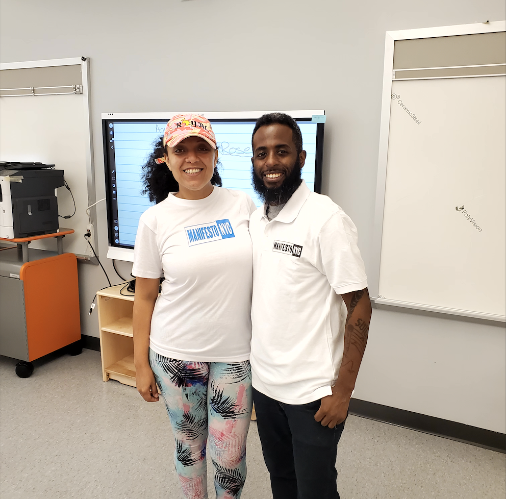 A woman and a man standing together in a classroom, both wearing white shirts with 'MANIFESTO NYC' printed on them, smiling at the camera.