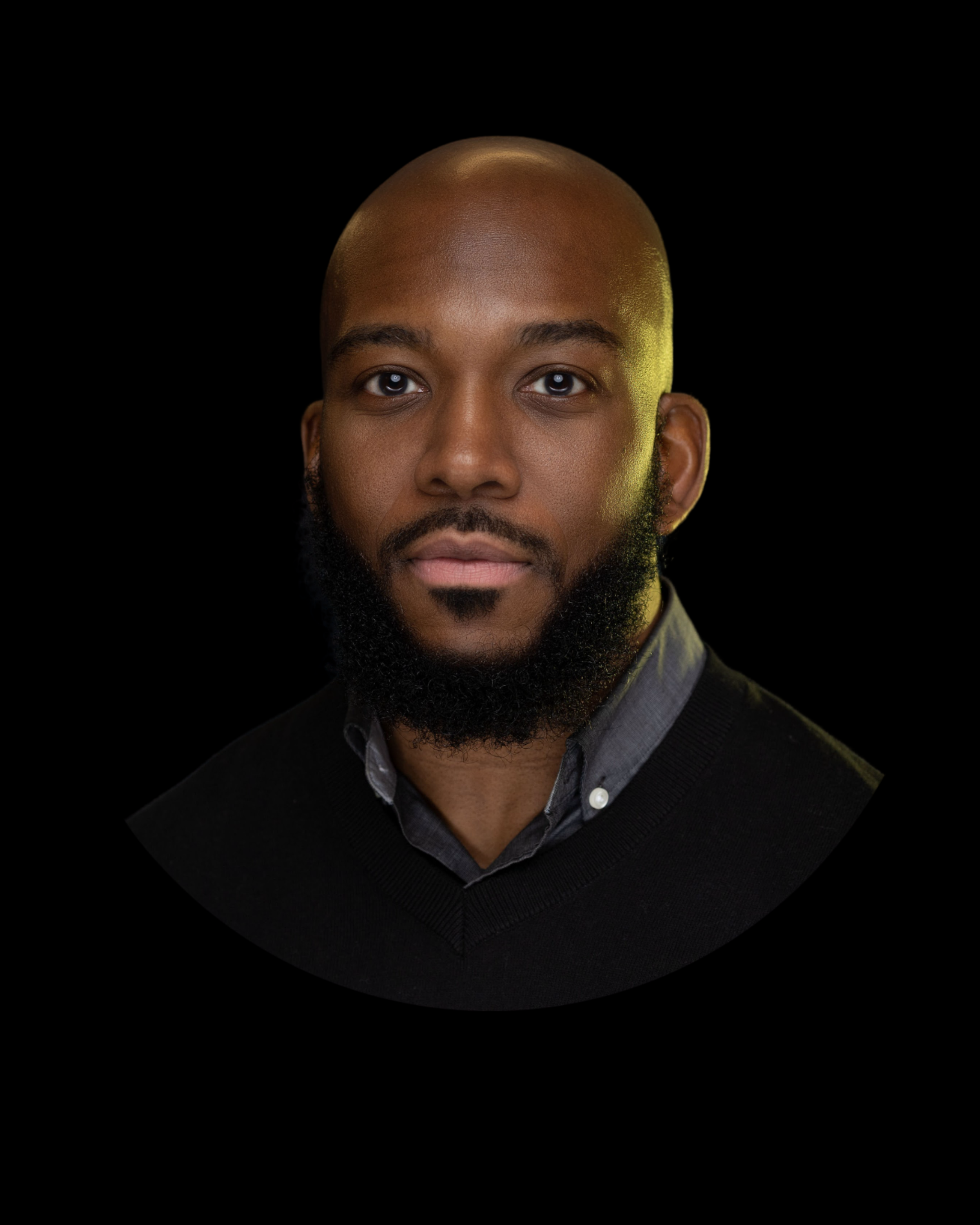 Close-up portrait of a Black man with a beard, wearing a collared shirt and a black sweater against a black background.