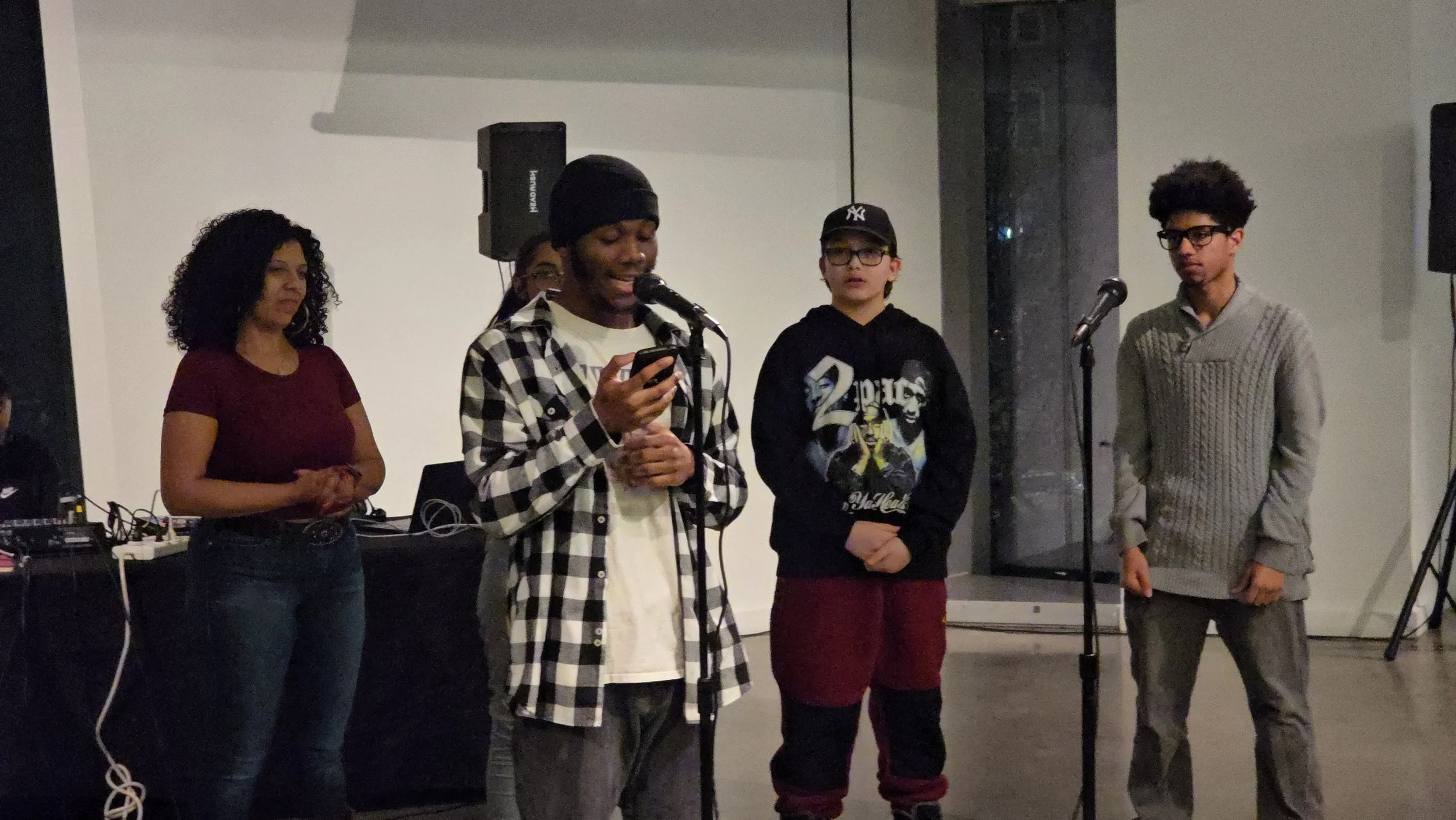 Group of five people on stage, including a man speaking into a microphone and three others listening, with audio equipment visible in the background.
