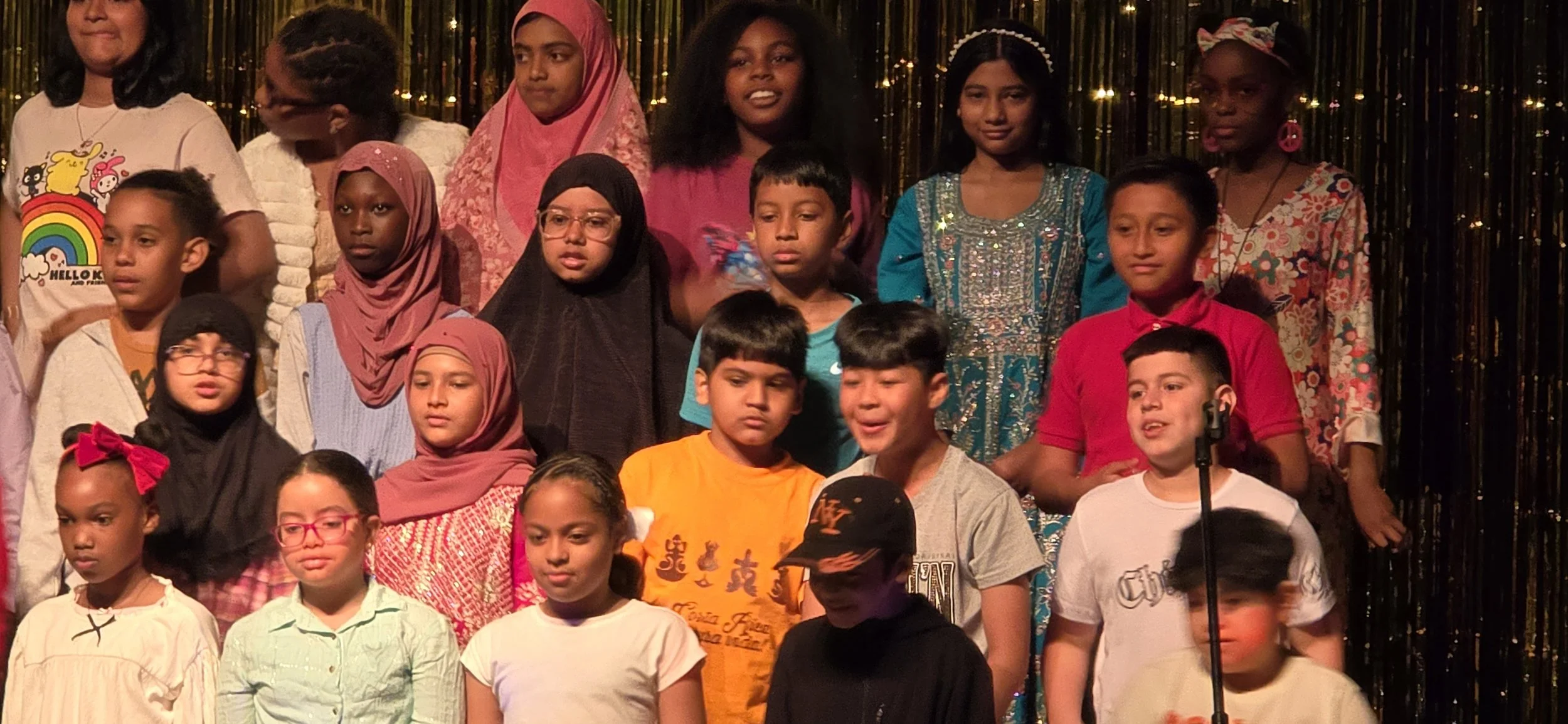 Group of children on stage during a school performance, with a gold curtain background.
