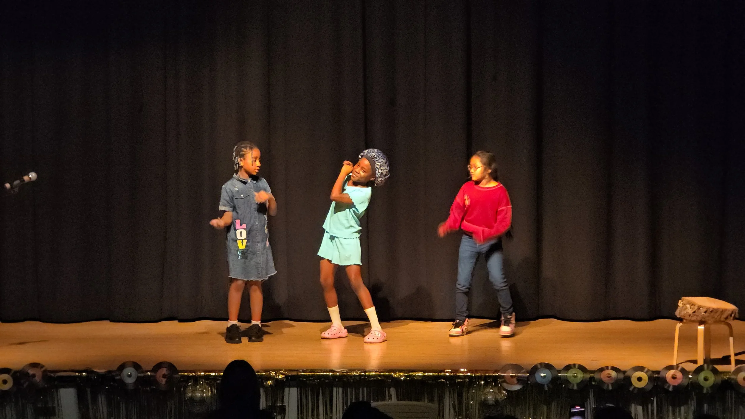 Three young girls performing on a stage with black curtains, one girl in the center wearing a blue outfit and pink Crocs, and another girl on the right wearing a red sweater and jeans, with a third girl on the left in a denim dress. A small wooden st