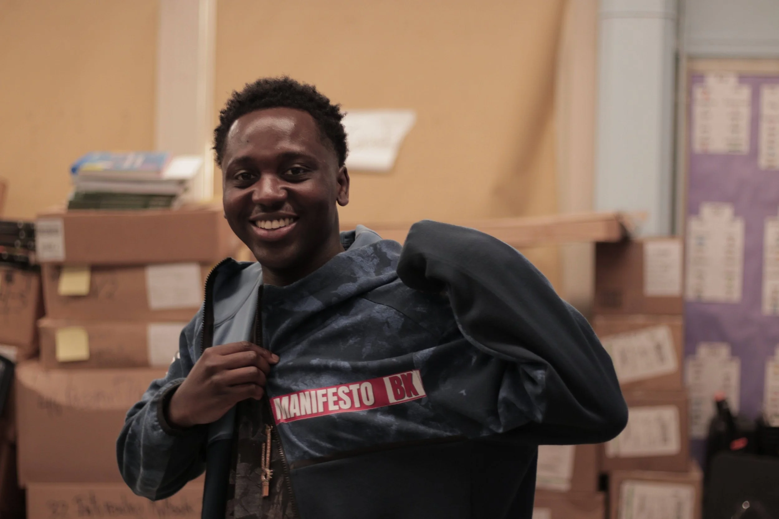 Young man smiling, holding jacket with 'MANIFESTO BK' patch, standing in storage room with boxes.
