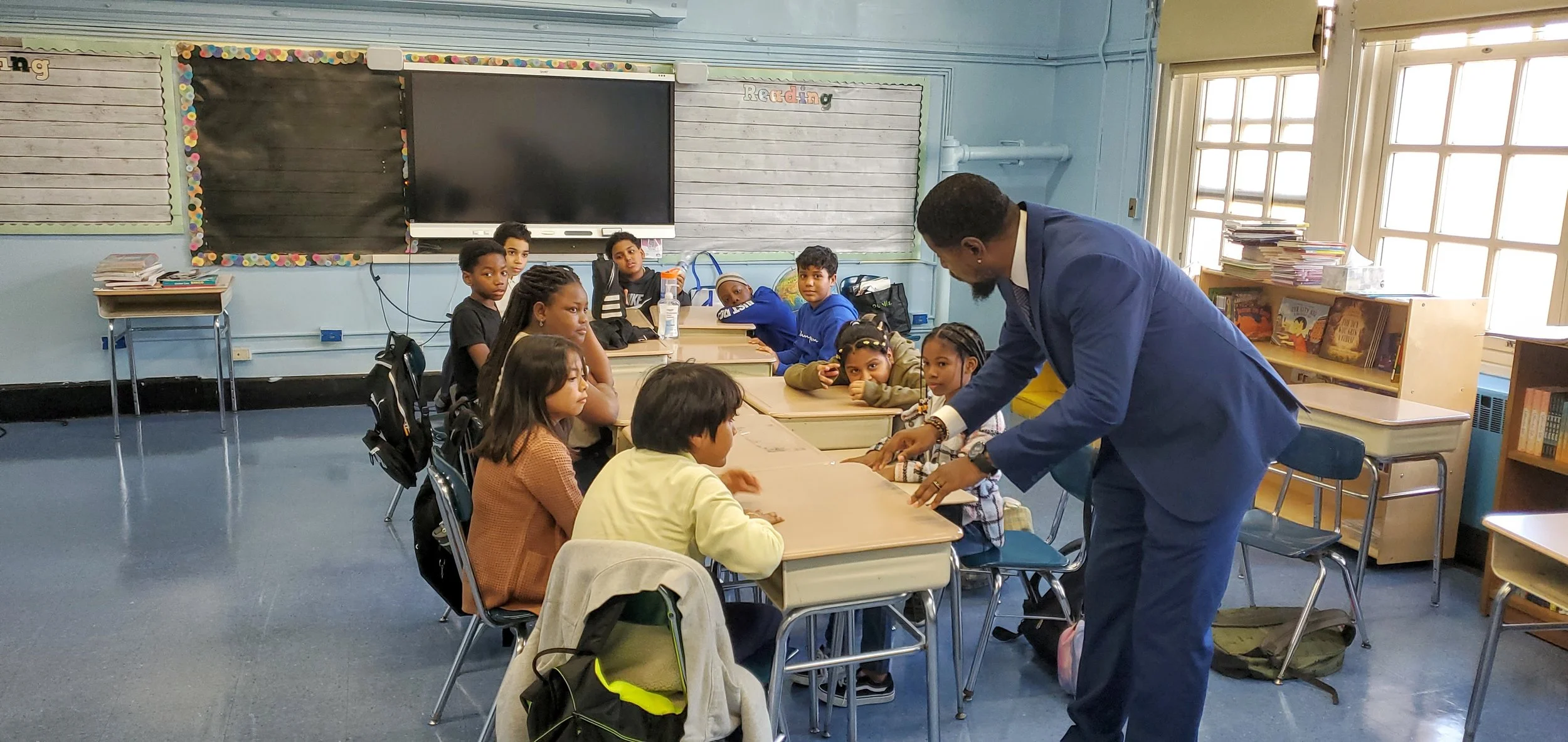 A classroom with a teacher in a blue suit engaging with students sitting at desks. The students watch the teacher attentively, and some have backpacks on their chairs. The classroom has large windows, a bookshelf with books, and a digital screen on t