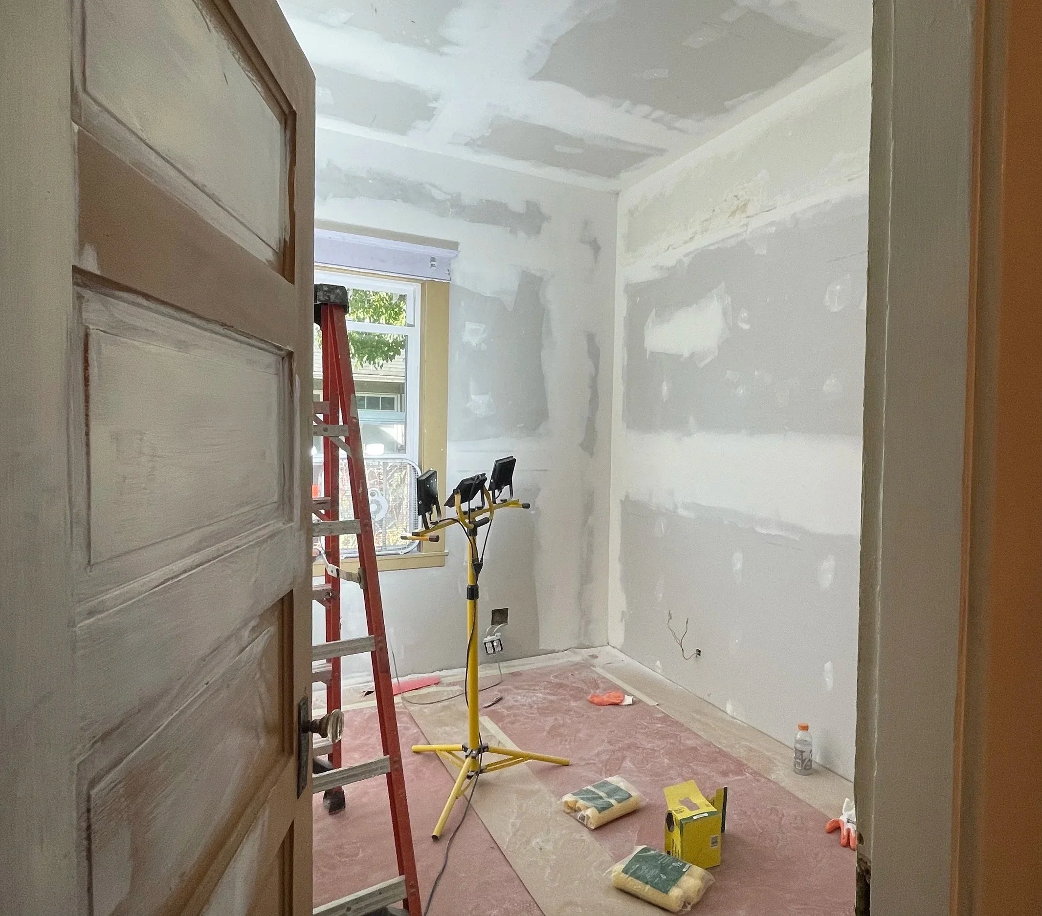 A room under renovation with drywall installation in progress, a window with light coming through, a red ladder, a yellow work light, and various tools and materials on the floor.