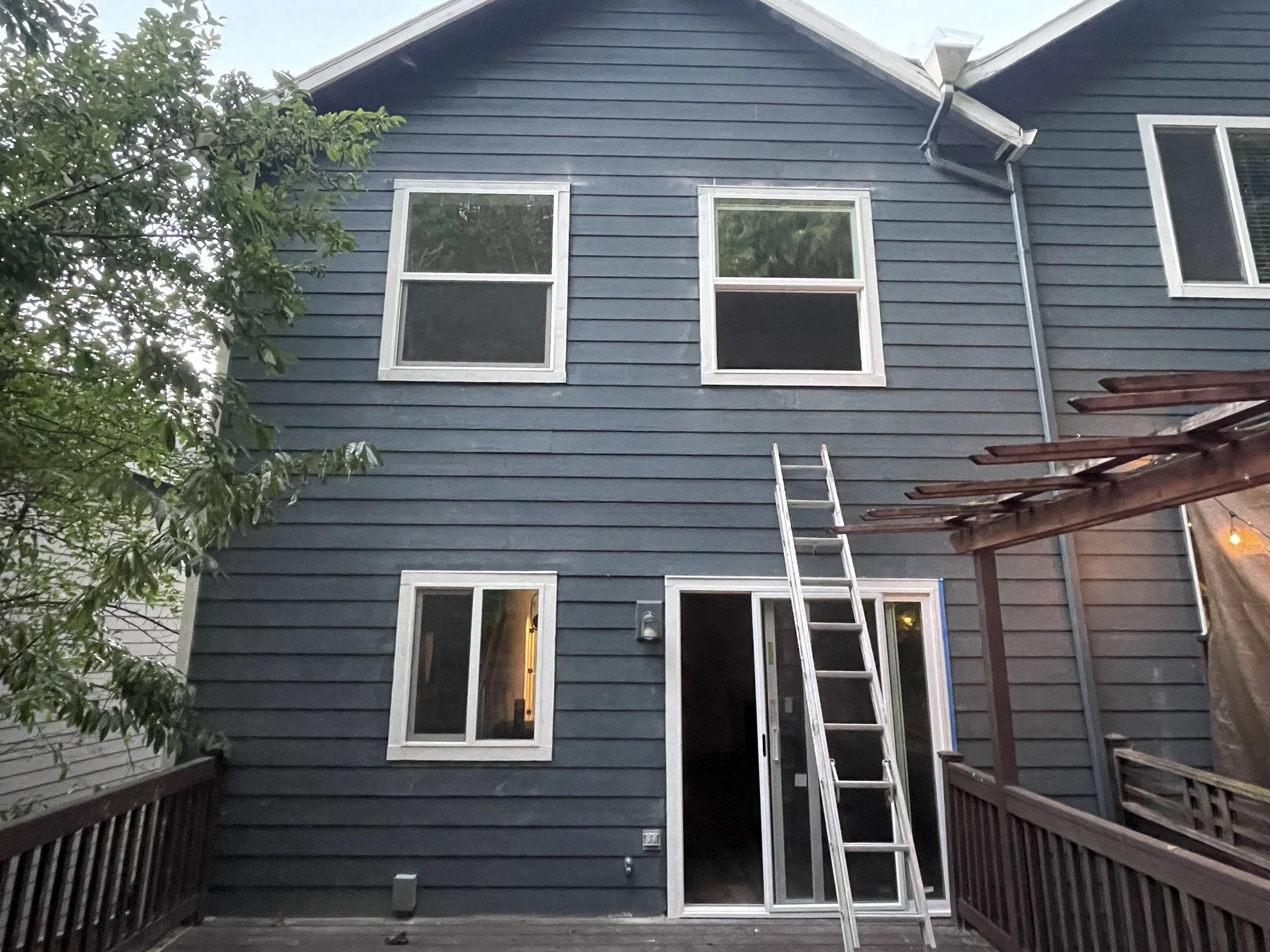 Rear view of a blue house with four windows, a sliding glass door, and a ladder leaning against the wall, with a deck in the foreground.
