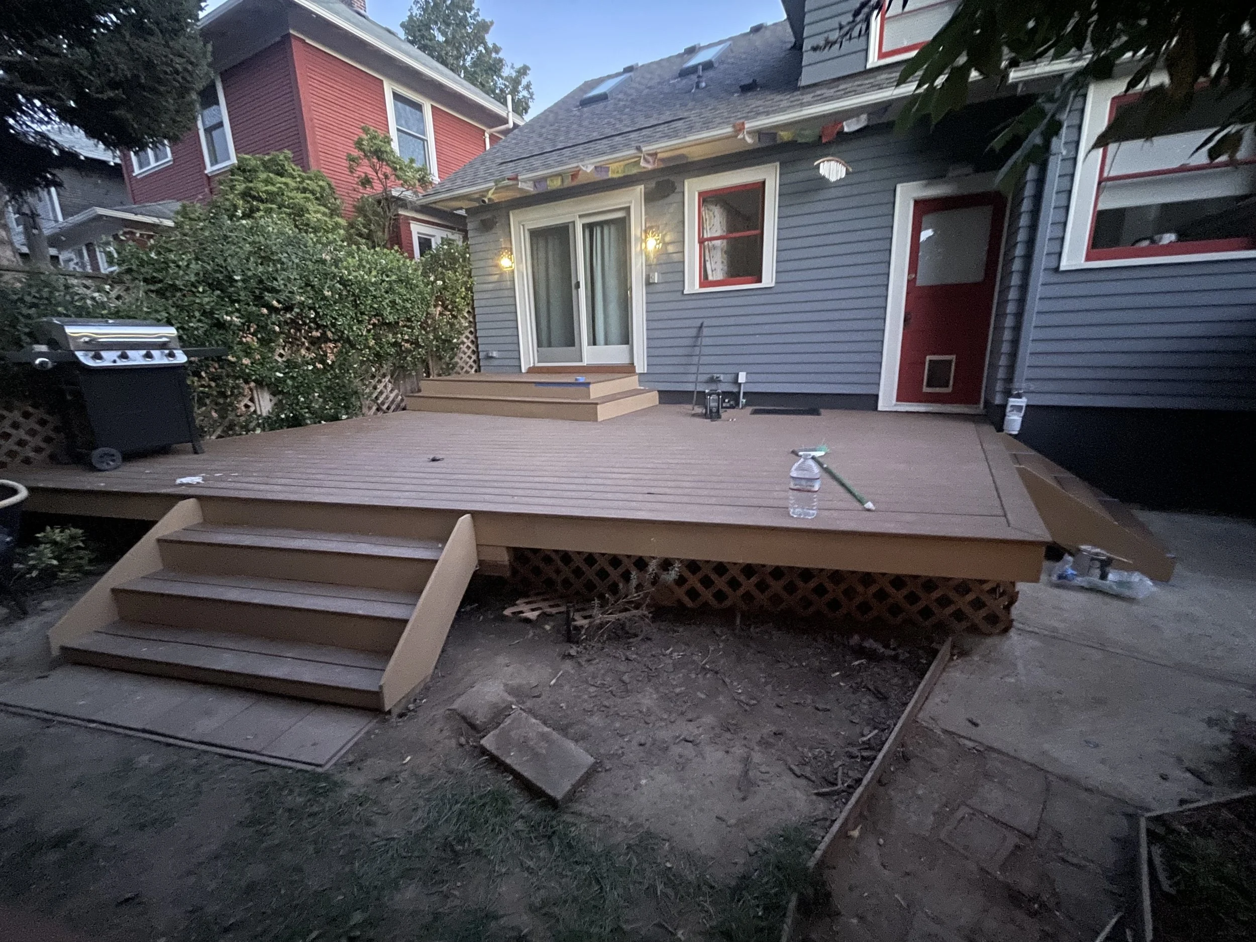 A newly built wooden deck with stairs attached to a gray house with red trim. The deck has a sliding glass door leading into the house. There are some gardening tools and a water bottle on the deck, and a grill on the side. The ground around the deck is still bare and under construction.