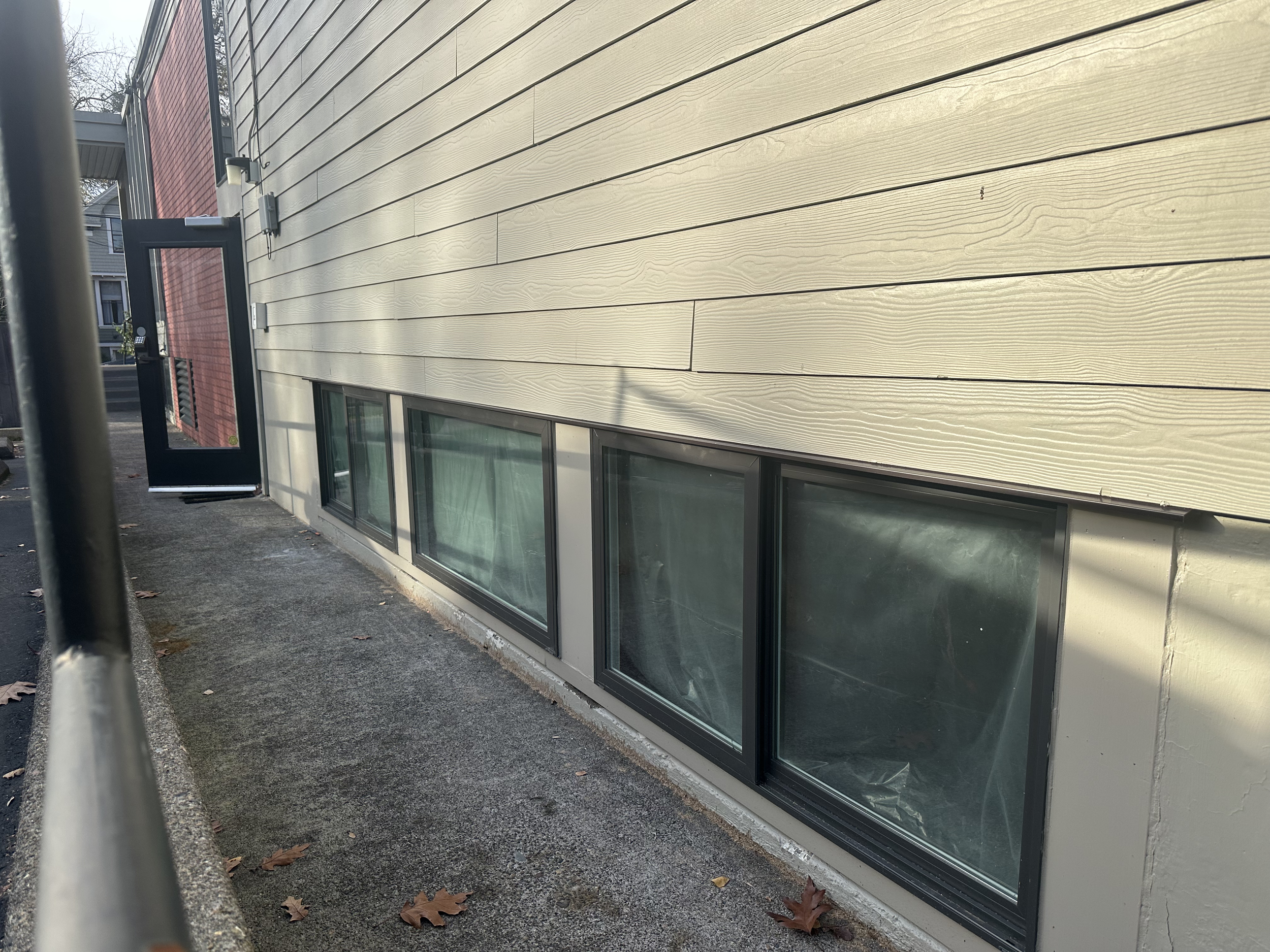 Exterior view of a building with horizontal beige siding, a row of sliding windows, and a black door with a glass panel, on a concrete sidewalk with fallen leaves.