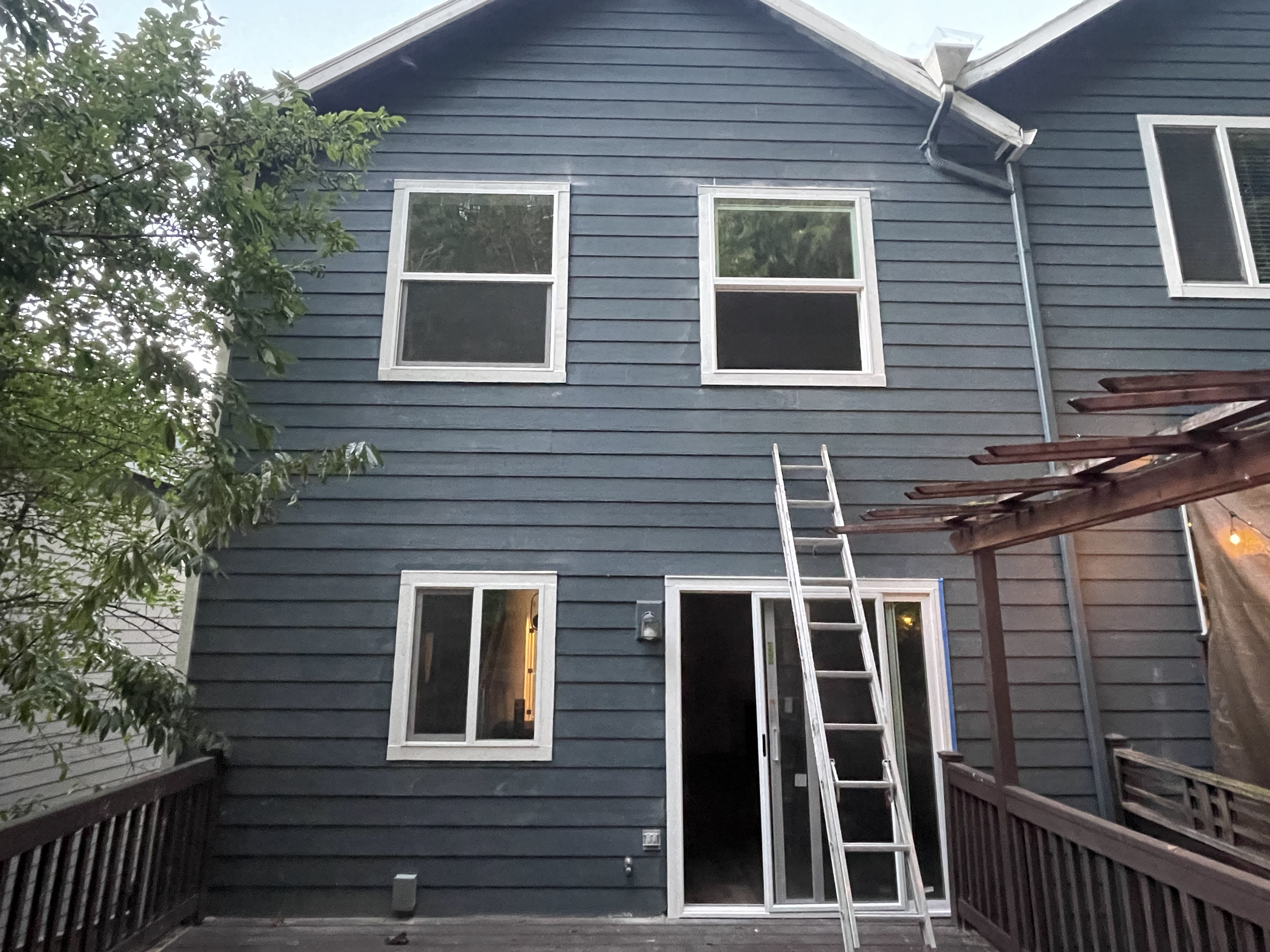 Back of a house with blue siding and four windows, a sliding door under repair with a ladder leaning against the house, and a wooden deck.