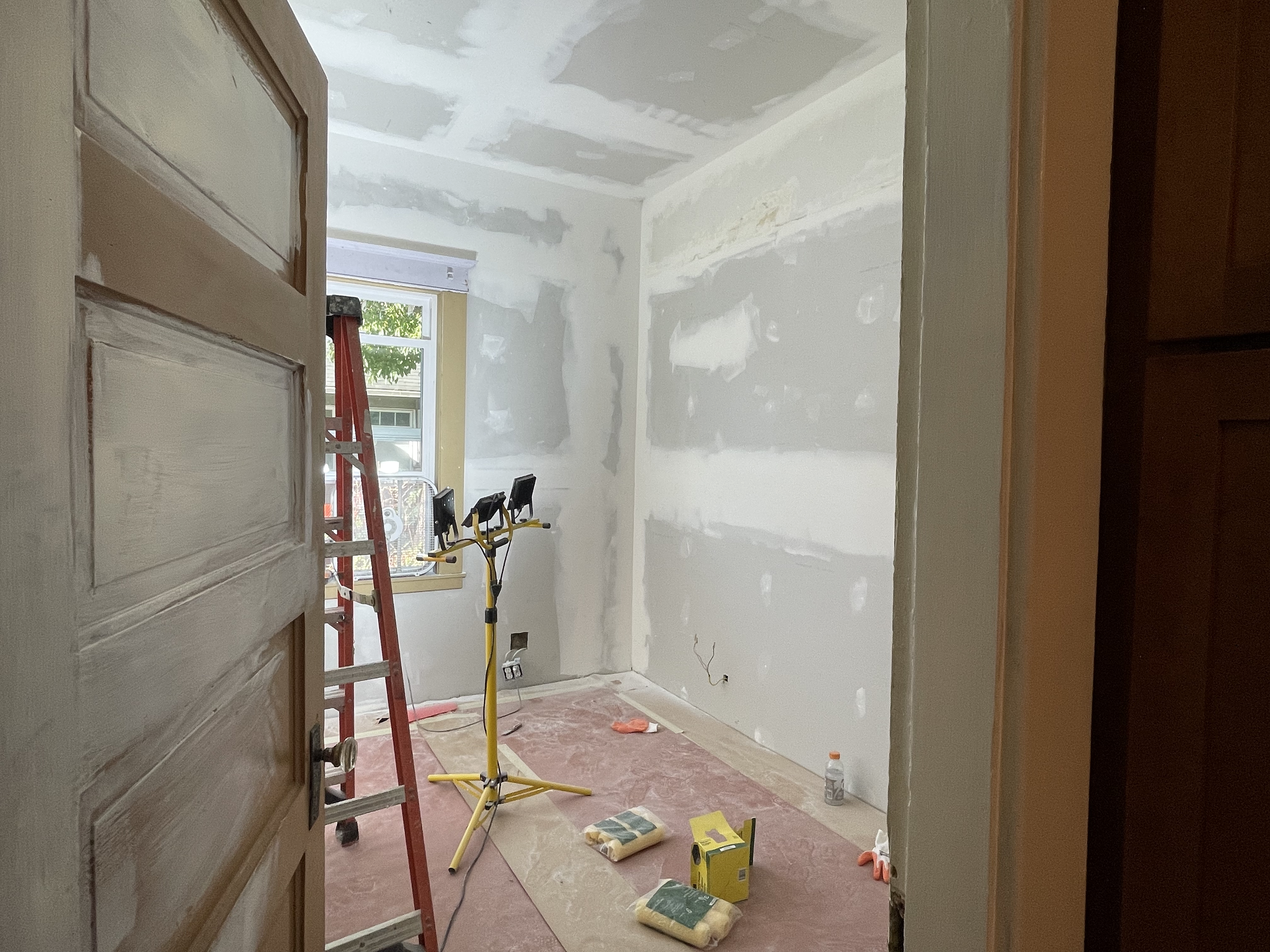 A small room under renovation with unfinished drywall, construction tools, a red ladder, and a light source coming from a window.
