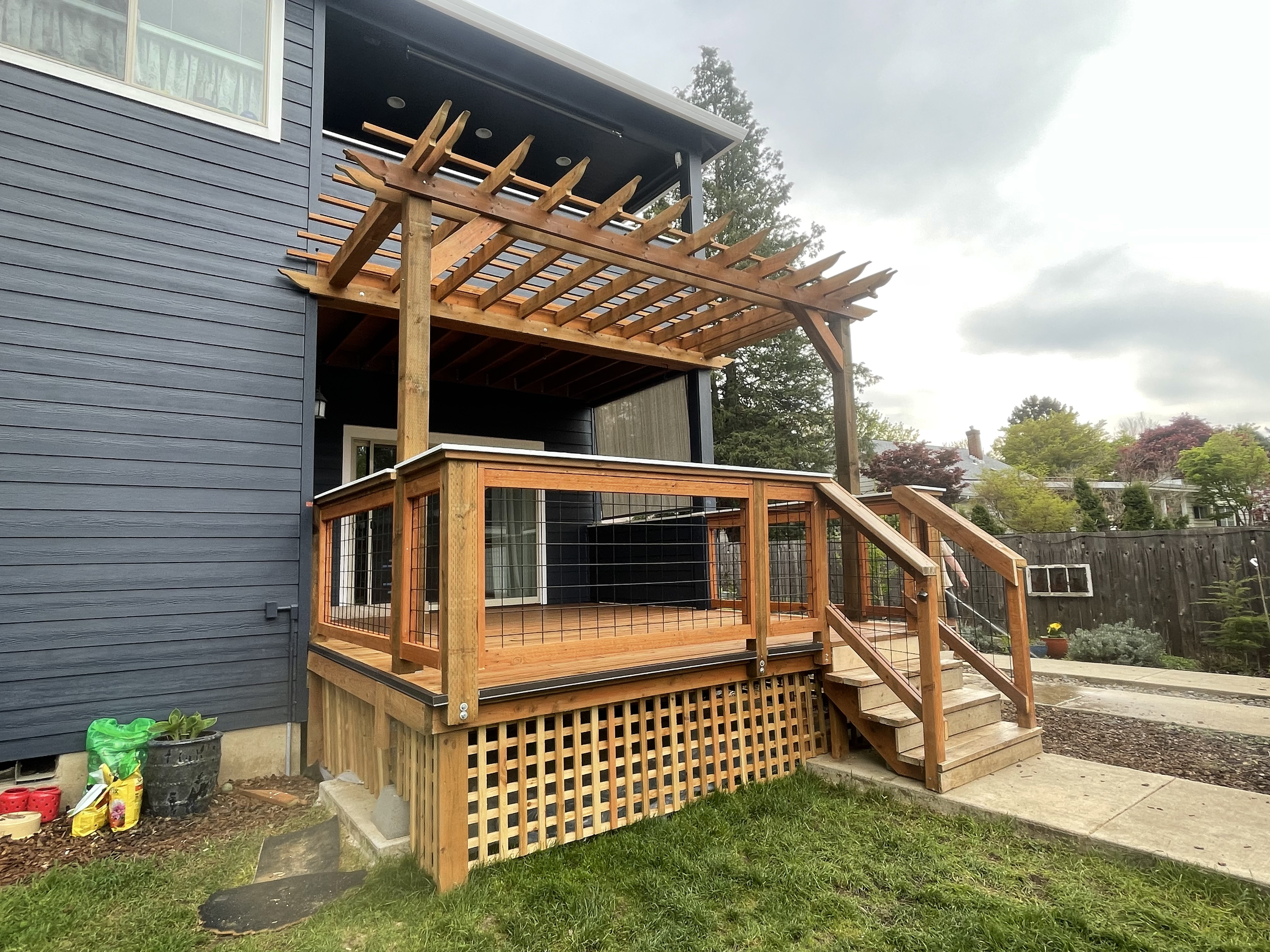 A wooden deck with stairs and a railing, attached to a dark blue house exterior. An unfinished pergola extends over the upper part of the deck, with a cloudy sky and trees in the background.