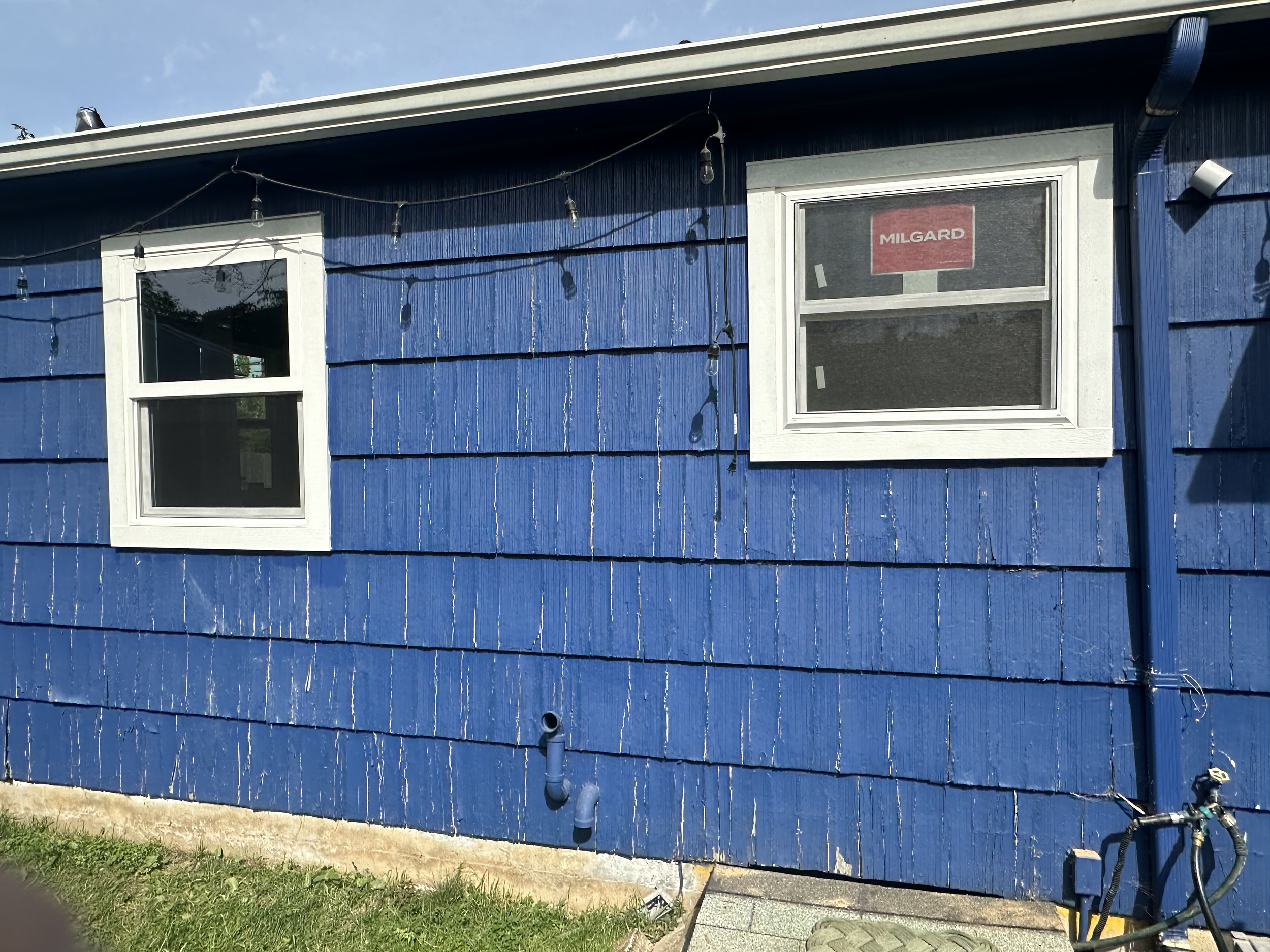Blue house with two white window frames and a gutter on the roof. String lights are hanging across the house. Grass and a sidewalk are in the foreground.