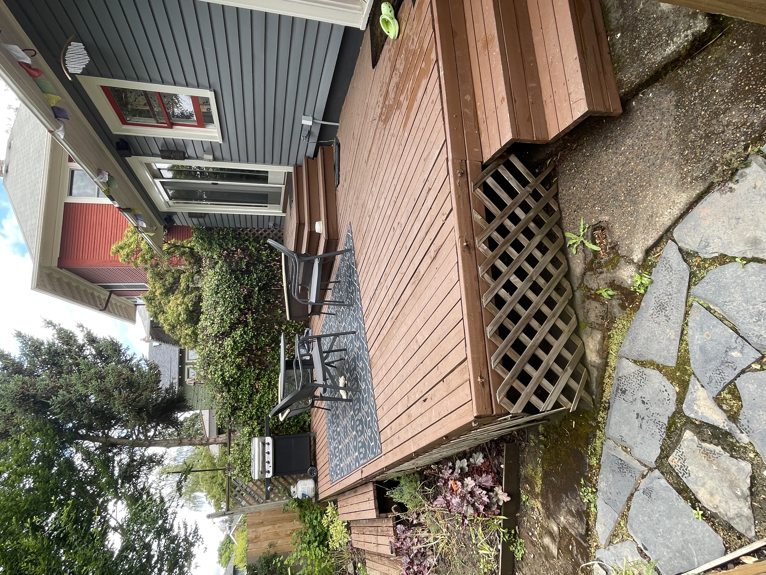 Backyard wooden deck with a small black table and four chairs, surrounded by plants and trees, adjacent to a house with gray siding and white trim, with a flag hanging on the house.