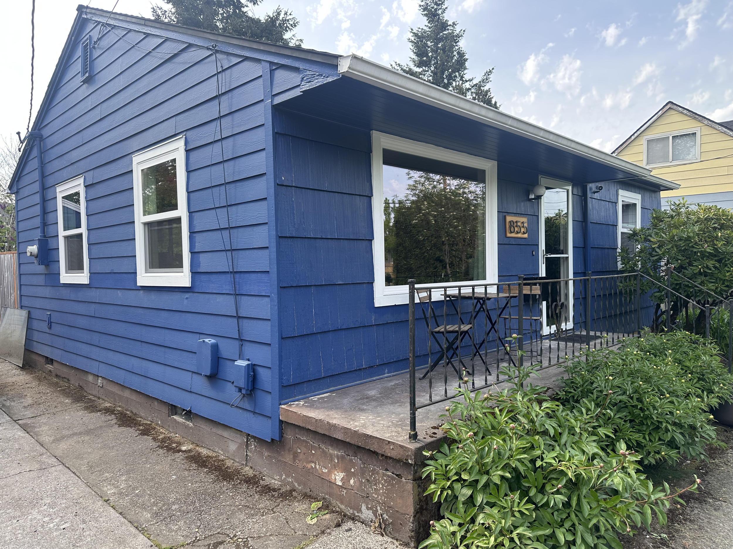 A blue house with white trim on the windows and a wooden sign with the house number 855 on it. The house has a small front porch with a black metal railing and is surrounded by greenery and a fence. The sky is partly cloudy.