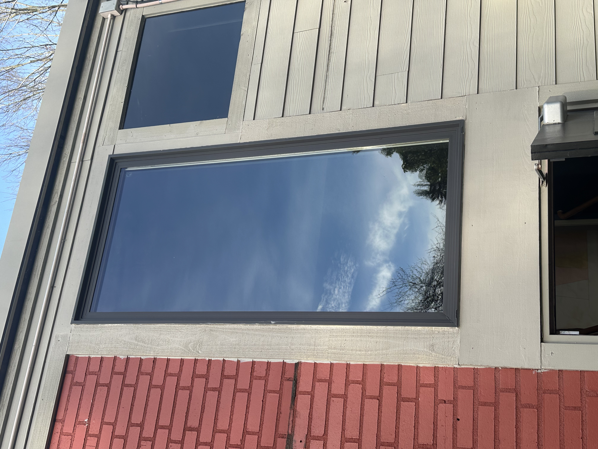 Reflection of clouds and trees in a large glass window on a building exterior with wood and brick siding.