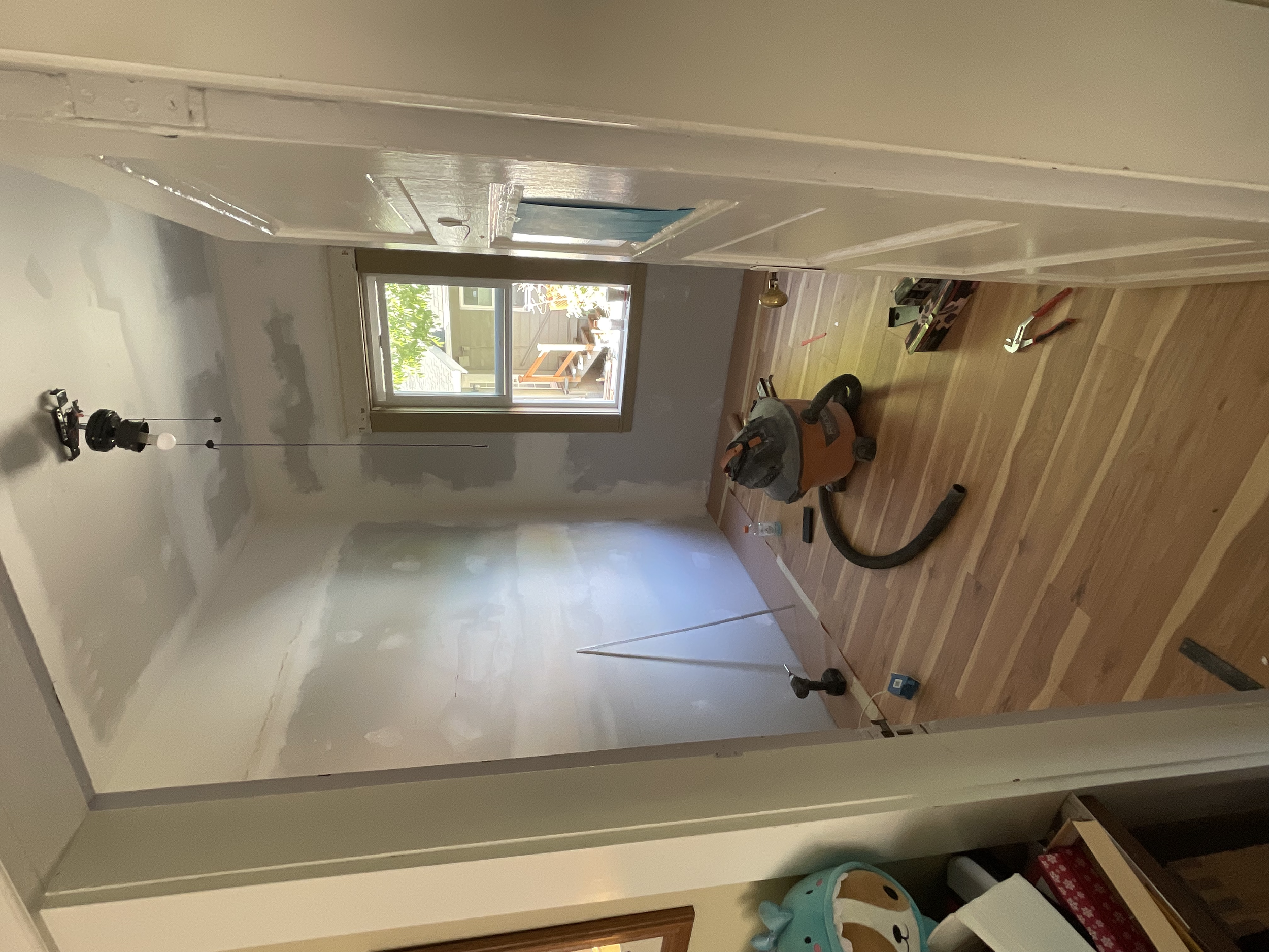 A room under renovation with drywall patches, a ceiling light fixture, and a window, containing a vacuum cleaner and construction tools on a hardwood floor.
