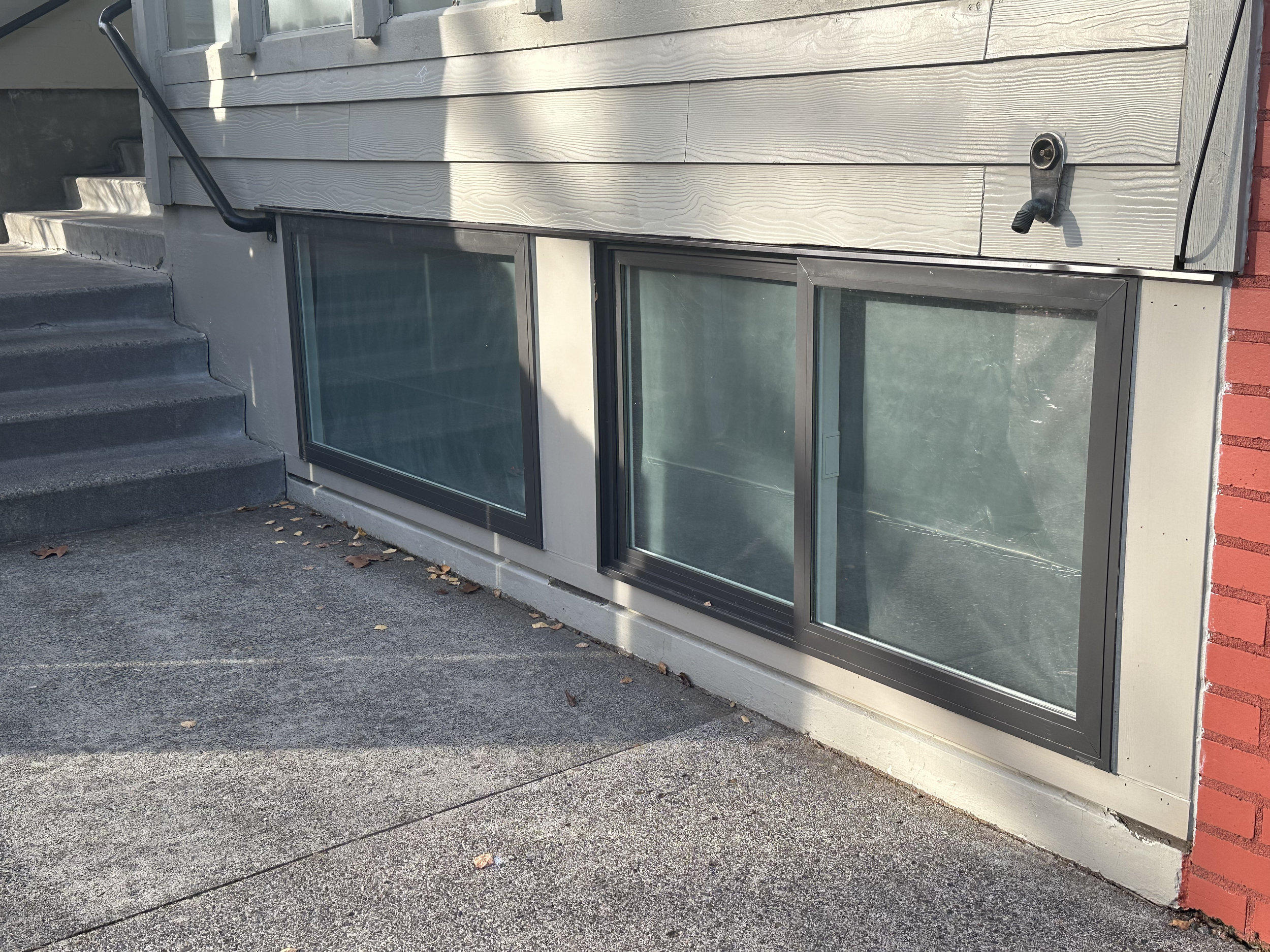 Basement window and concrete stairs outside of a house with gray siding and red brick corner.