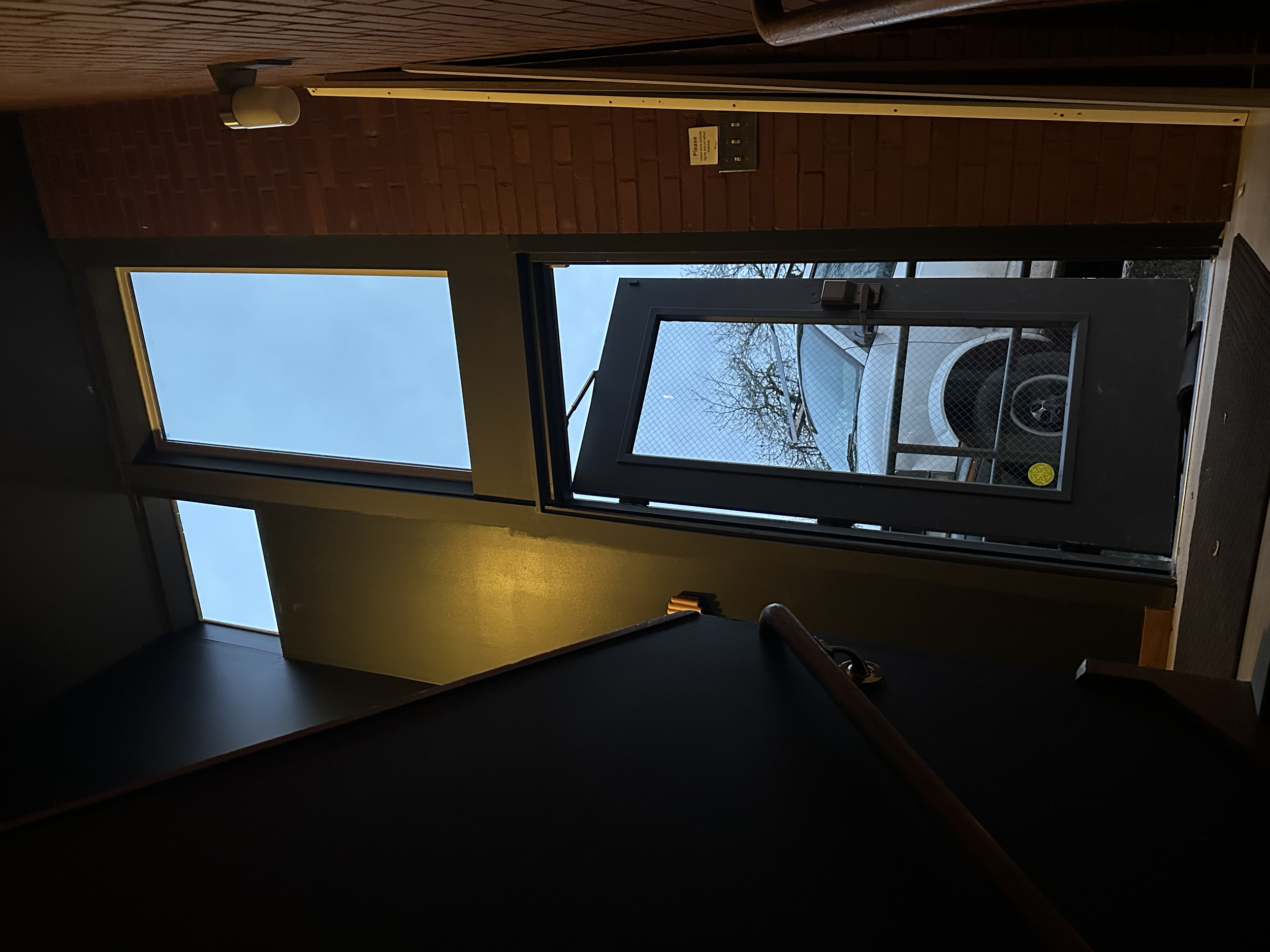 Indoor view showing a door with a large glass window and a small open window above, leading outside to a parking lot with a white vehicle, brick and painted walls, and a staircase railing visible.