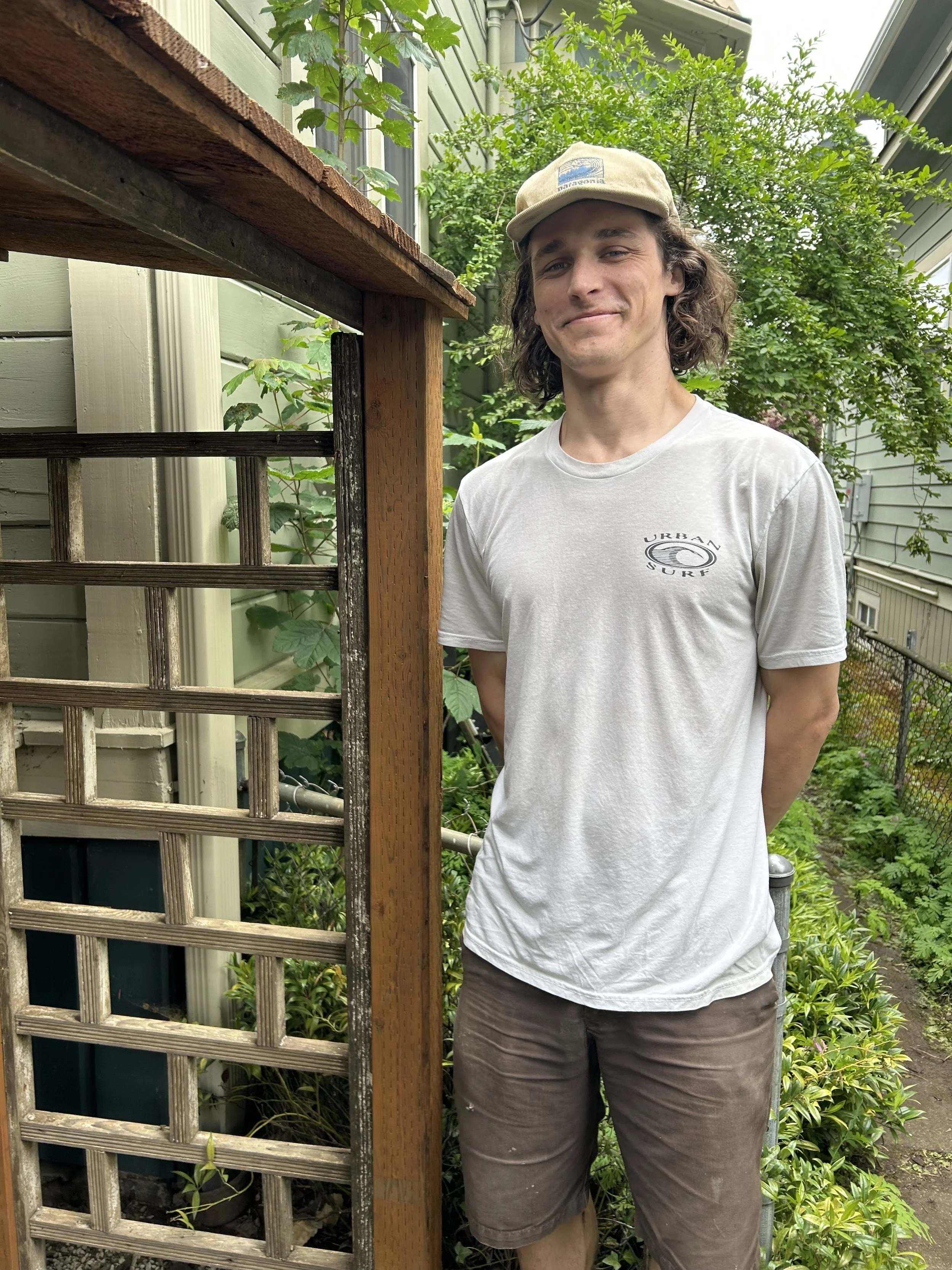 Young man standing outdoors next to a garden trellis with lush green plants in the background, wearing a light gray t-shirt, brown shorts, and a beige cap.