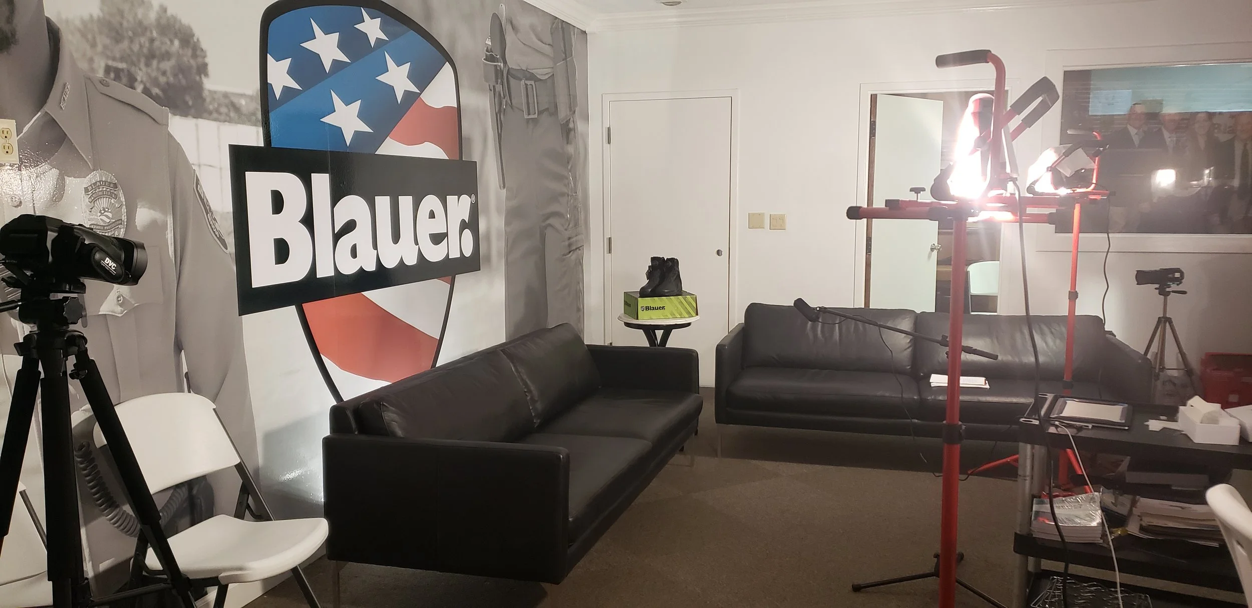 A room set up for filming or streaming, with a large 'Blauer' sign on the wall, a police uniform on display, two black leather couches, a white chair, professional lighting equipment, and a window showing a blurred video screen.